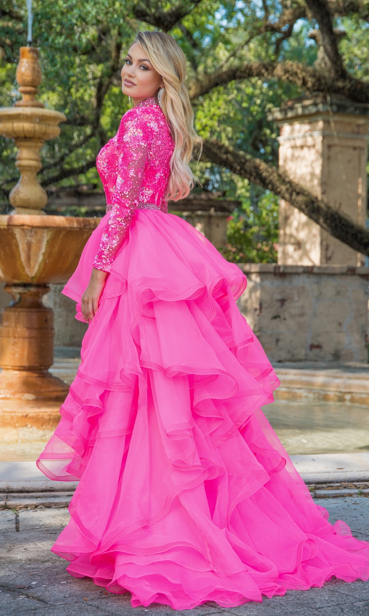 A woman stands outdoors by a fountain, wearing the Ava Presley 28595 Ruffle Short Sequin Romper in bright pink with long sleeves and a dramatic ruffled overskirt for elegance.