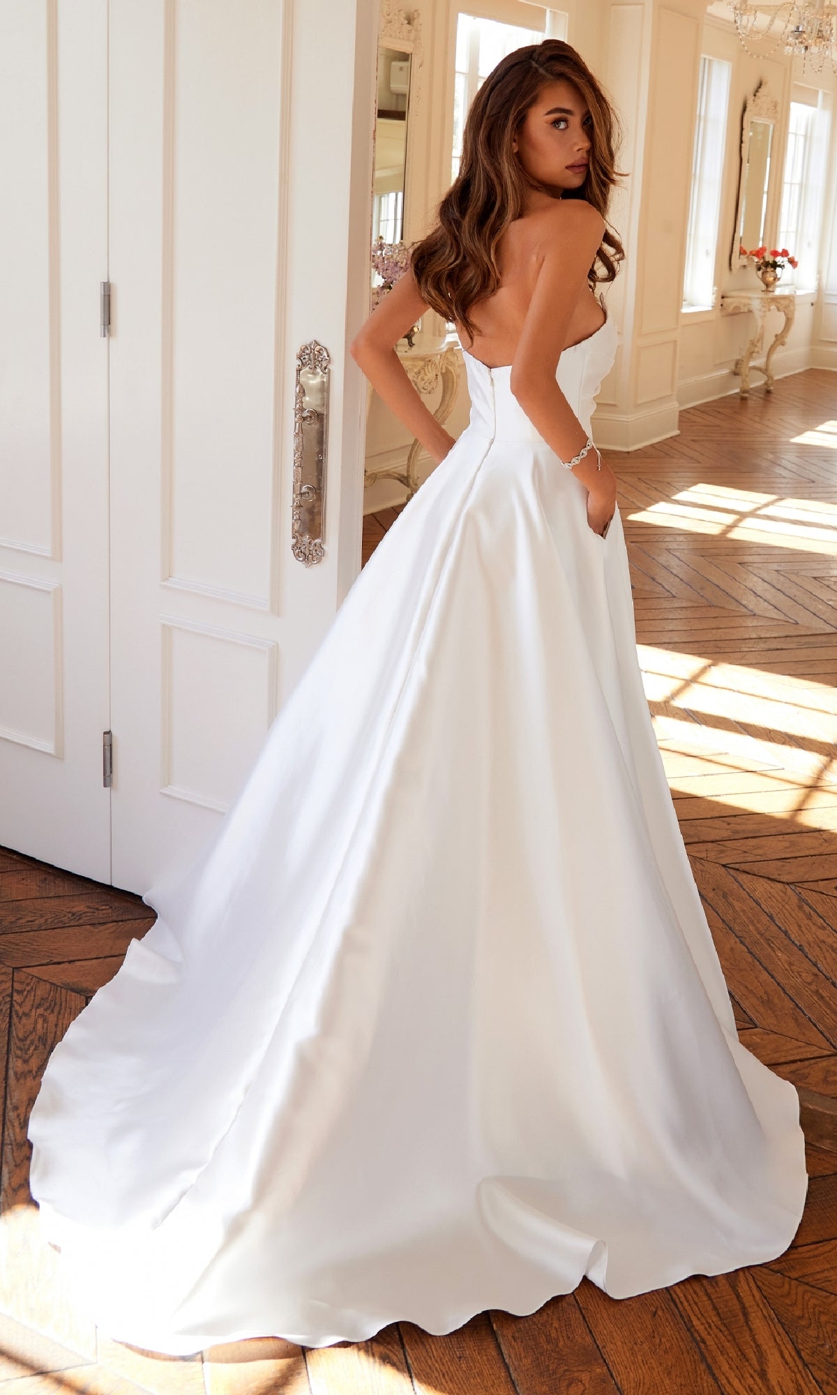 A woman stands indoors wearing the Long Prom Dress: Juliet JT2885CW, a strapless gown with a long train, looking over her shoulder.