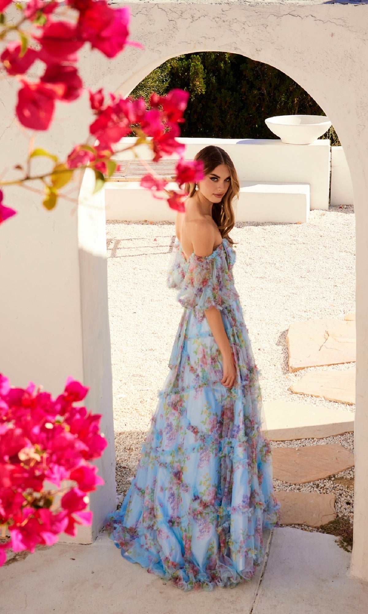 A woman in the Off-Shoulder Long Print Prom Dress: Juliet JT2887C stands outdoors beneath a white archway, with bright pink flowers in the foreground.