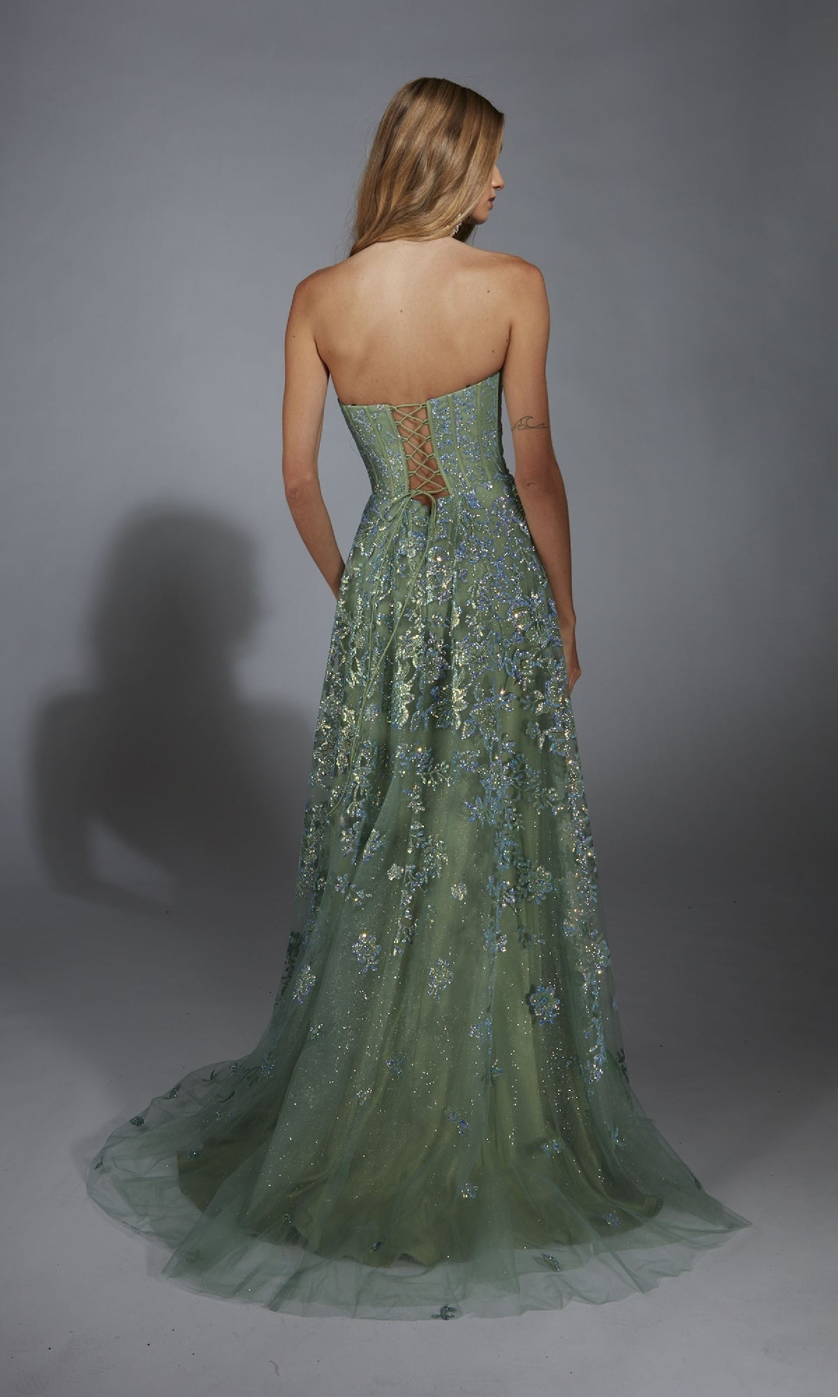 A woman faces away, wearing the Long Green Prom Dress: Aerie Couture 288, which features a strapless corset bodice and sheer tulle skirt, set against a plain gray background.