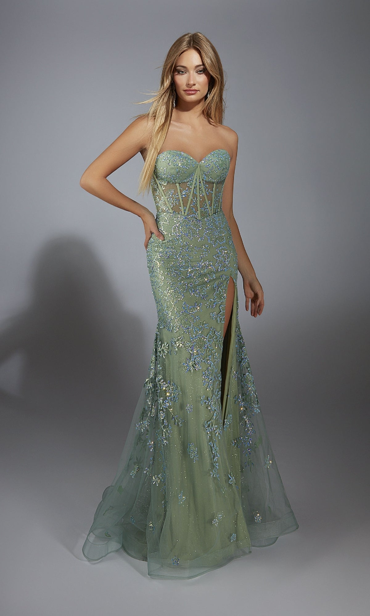 A woman wearing the Long Green Glitter Prom Dress: Aerie Couture 289, a strapless mint green gown with floral embellishments, corset bodice, sheer details, and a thigh-high slit, stands against a plain gray background.