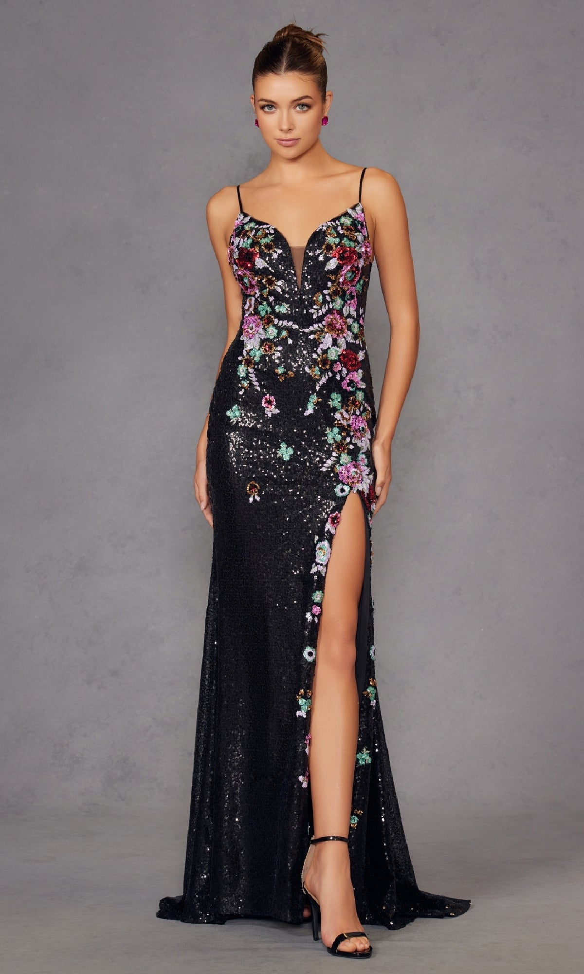 A woman models the Juliet JT2909K, a long black sequin floral prom gown with a deep v-neck, spaghetti straps, and high slit, standing against a plain gray background.