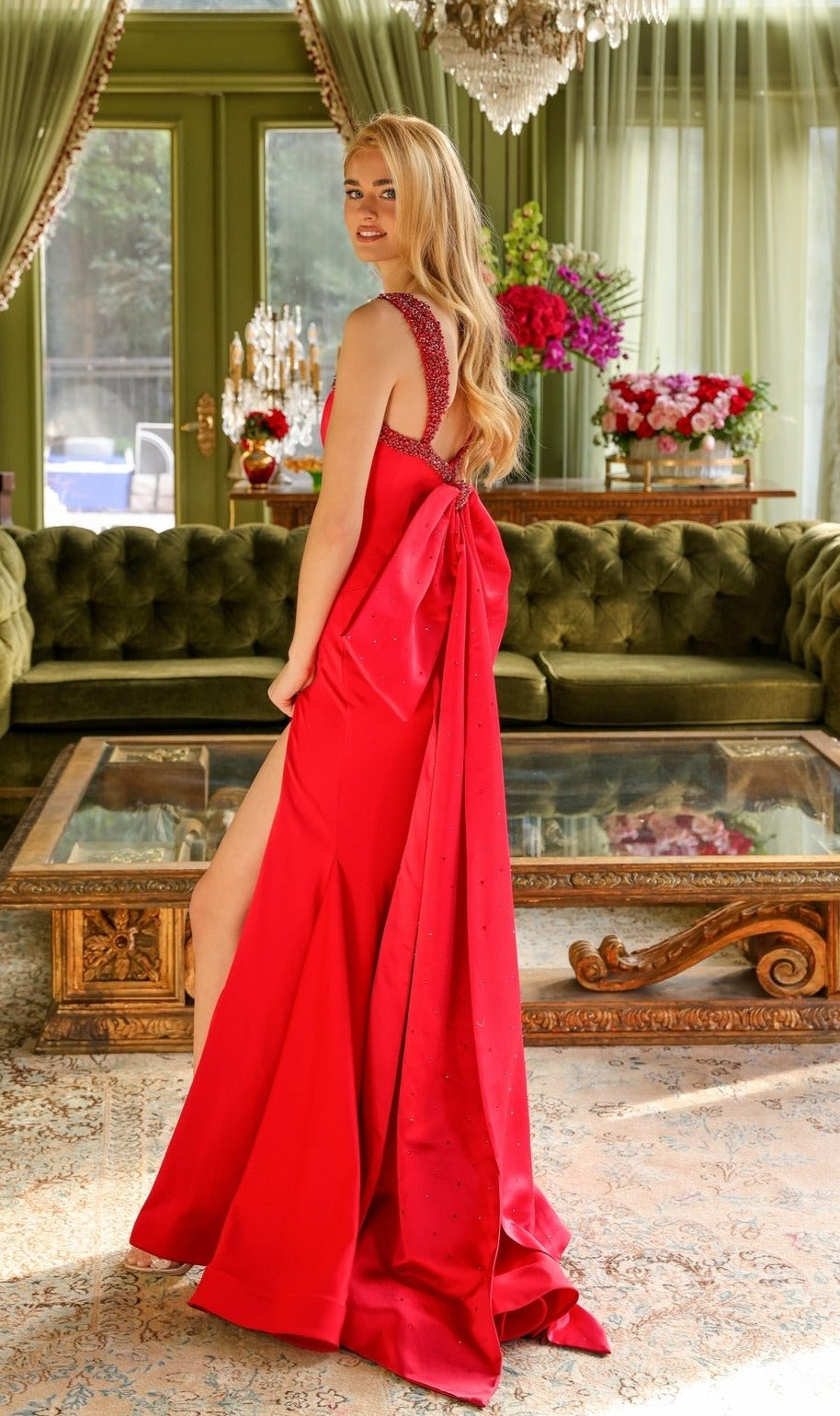 A woman wears the Long Prom Dress 29121 by Ava Presley, a fitted red gown with a bow back, as she stands in a decorated room with green sofas, a glass table, and floral arrangements.