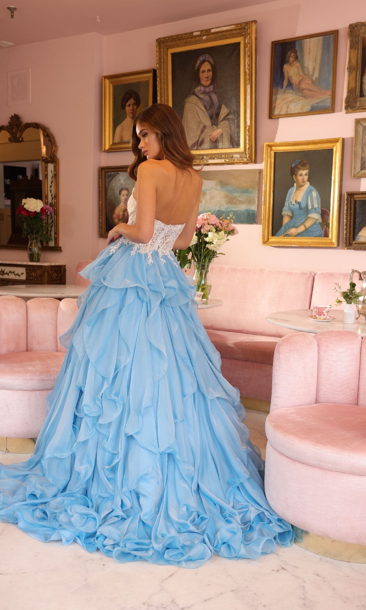 A woman wearing the Ava Presley 29133 strapless ruffle ball gown stands in a pink room decorated with vintage portraits and floral arrangements.