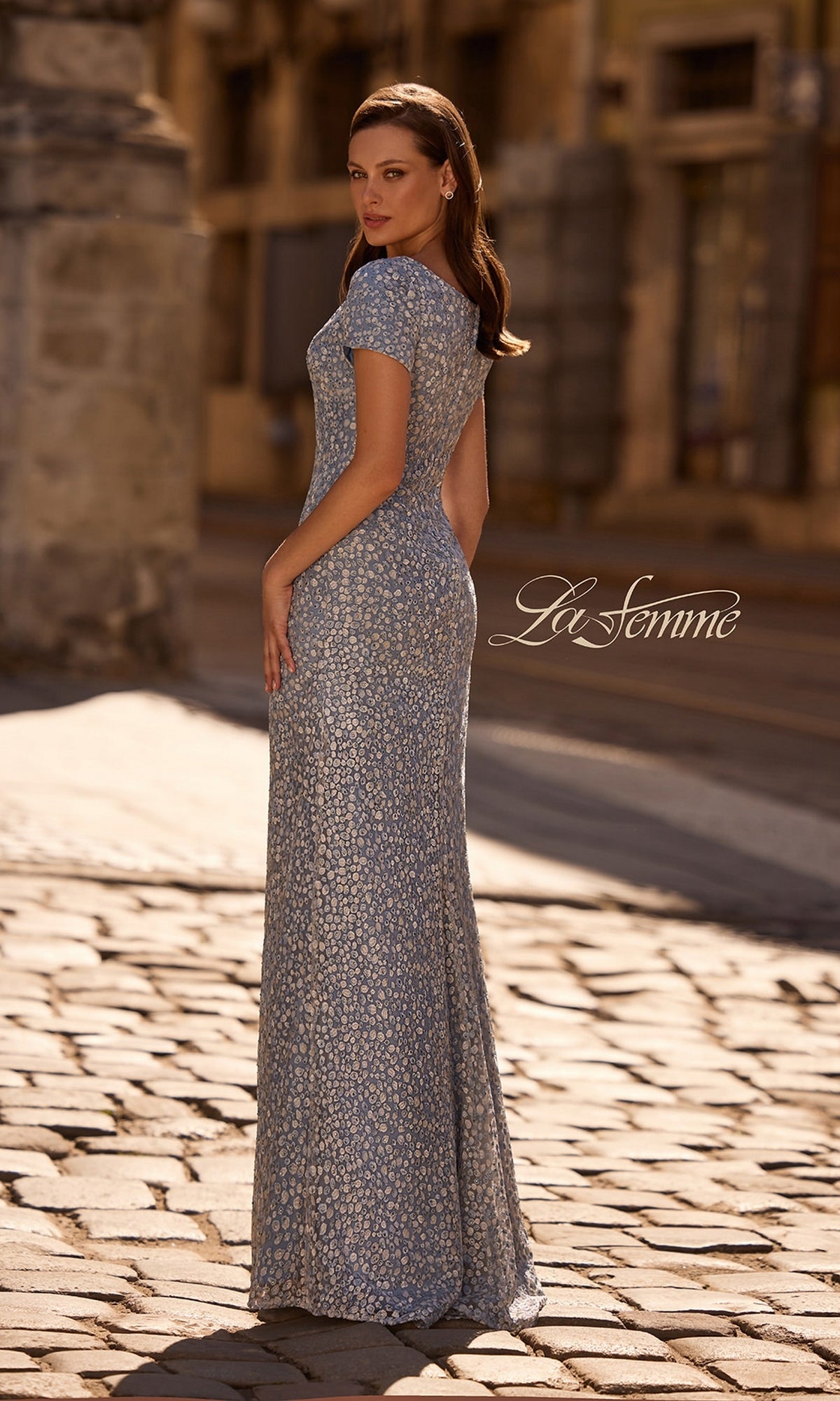 A woman in a silver sequin sheath stands on a cobblestone street in the Long Formal Dress: La Femme 29134, glancing over her shoulder. The “La Femme” logo appears on the right.