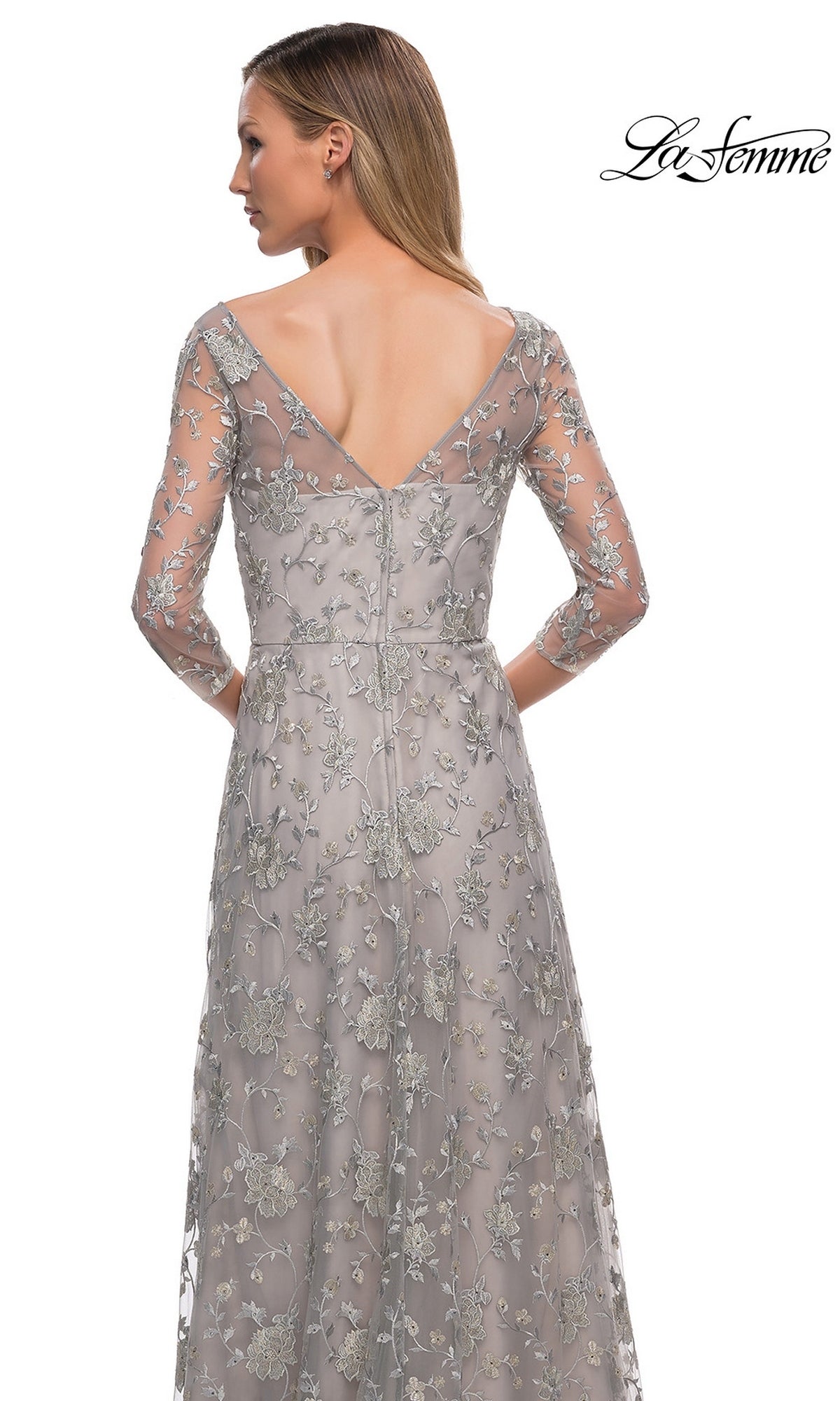 Woman wearing Long Formal Dress: La Femme 29153—a long, silver gown with sheer floral lace-embroidery and deep V-back, standing facing away.
