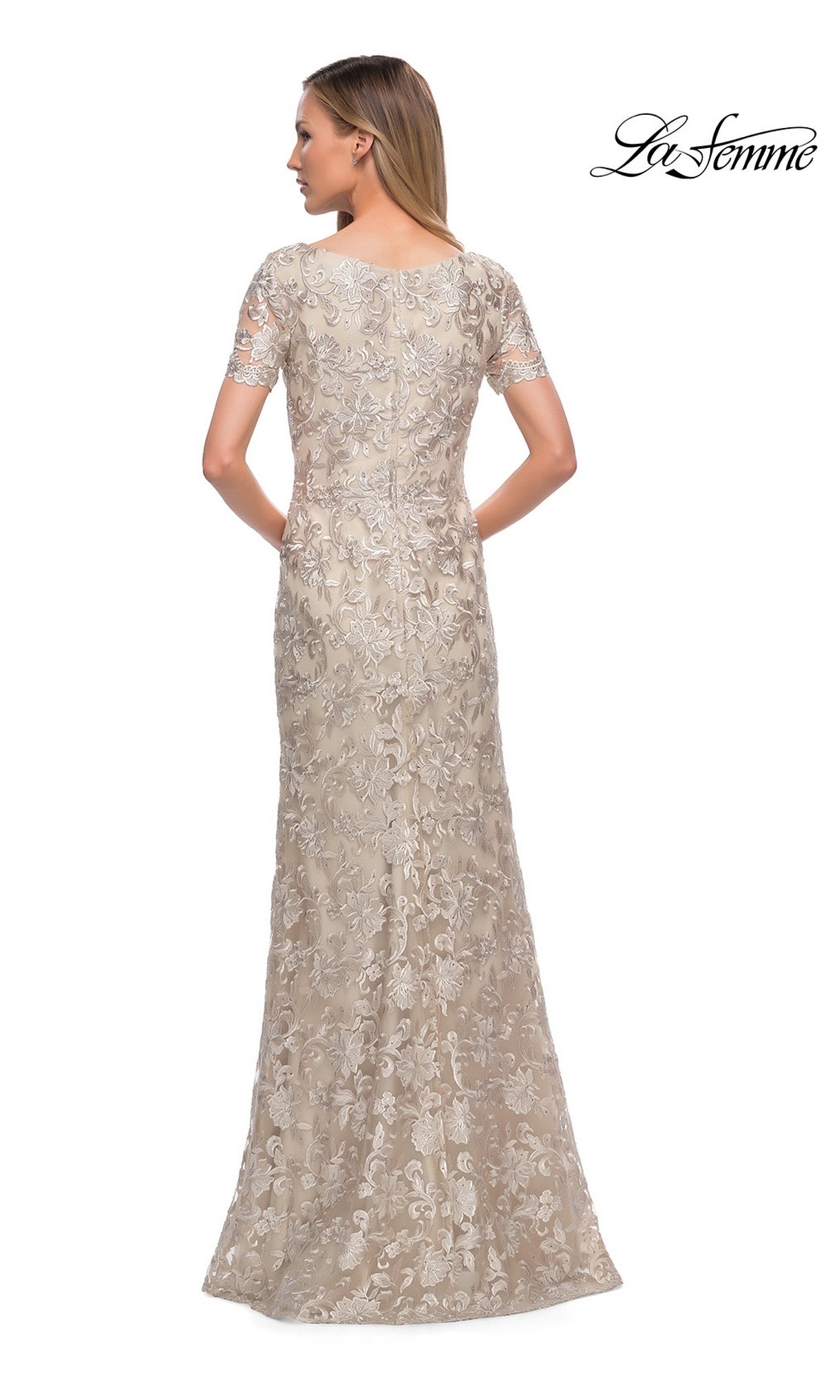 A woman is shown from the back wearing the La Femme 29161, a long formal, champagne-colored lace sheath dress with short sleeves and intricate floral embroidery. The “La Femme” logo appears in the top right corner. This elegant floor-length dress is an ideal choice for a mother-of-the-bride ensemble.