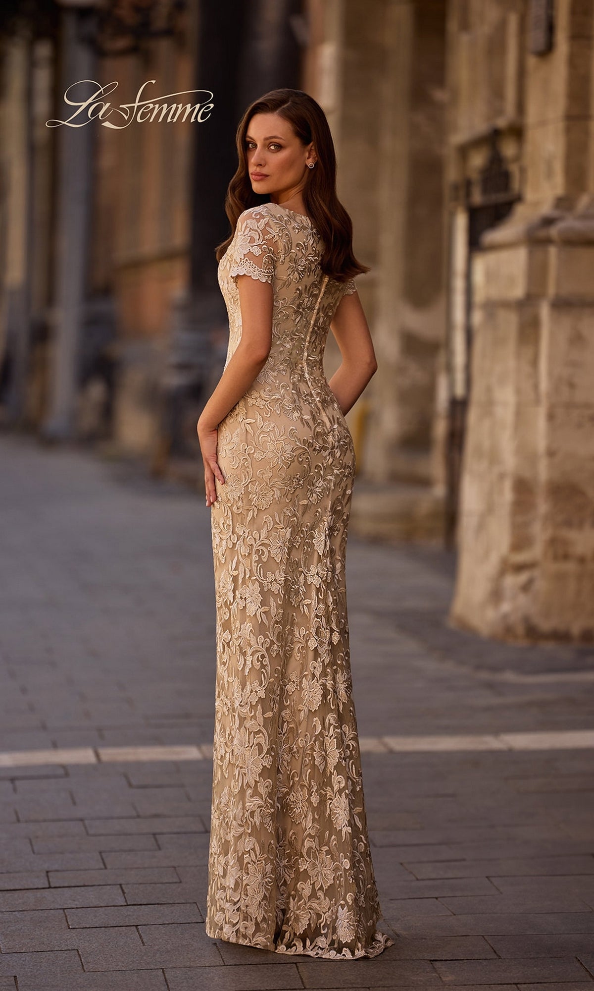 A woman wearing the La Femme 29161 long formal dress stands on a cobblestone street facing away, with historic buildings in the background.