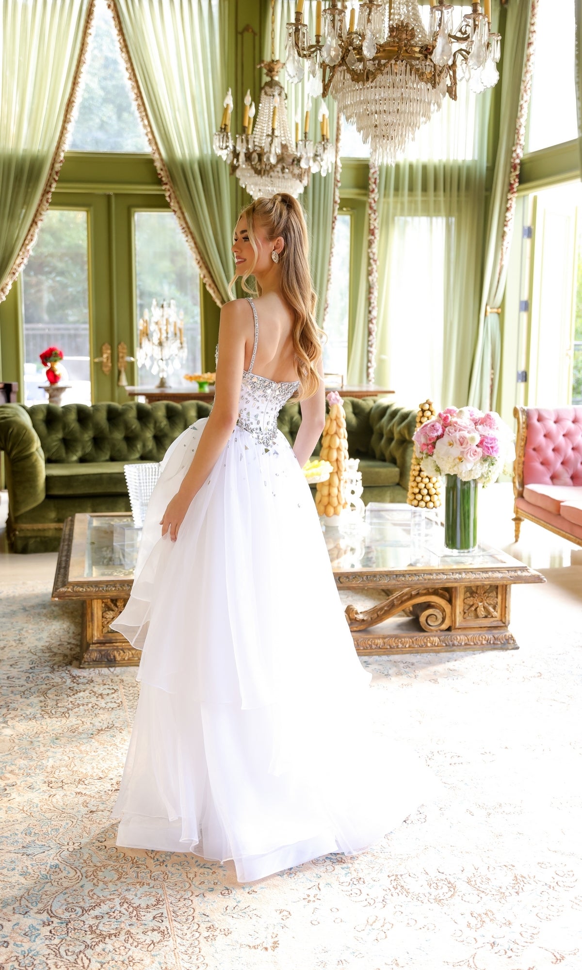 A woman wearing the Ava Presley 29189 Tiered Long Prom Ball Gown stands in an elegant room with green curtains, chandeliers, and ornate furniture, looking over her shoulder and smiling.