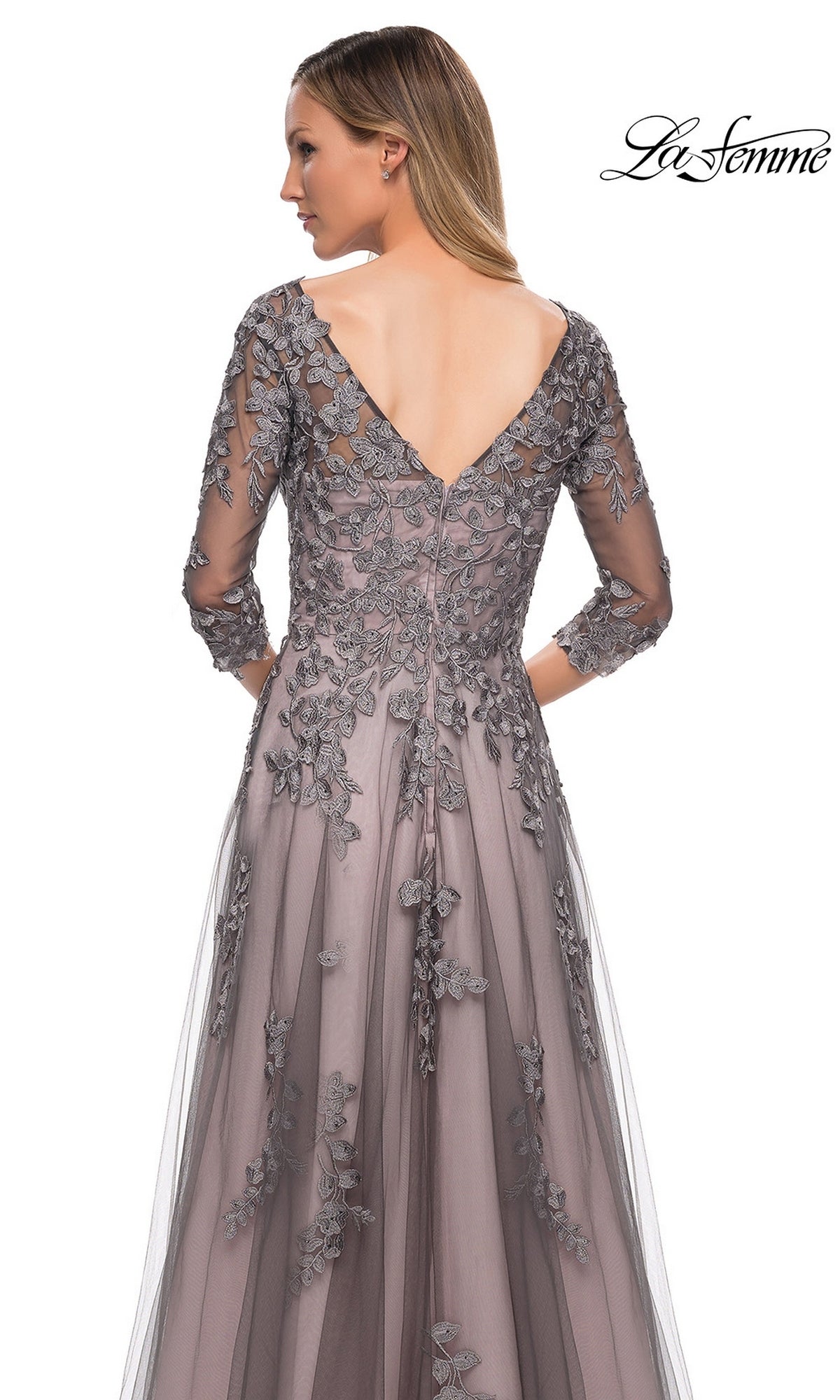 A woman stands facing away from the camera, elegantly showcasing the delicate details of the La Femme 29205 long formal dress—an A-line tulle gown in gray with sheer sleeves, intricate embroidery, and a V-shaped back.