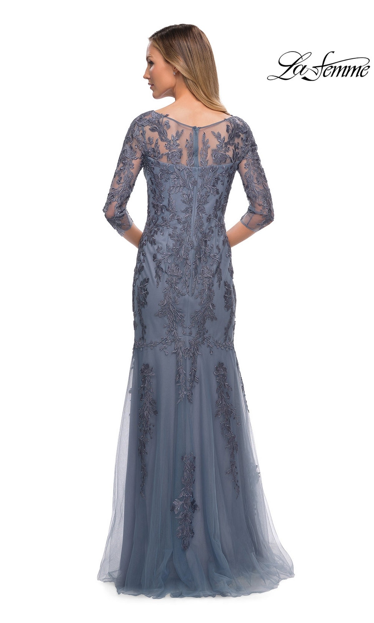 Woman in Long Formal Dress: La Femme 29226, blue lace with sheer sleeves and floral embroidery, standing with back to camera. “La Femme” logo top right.