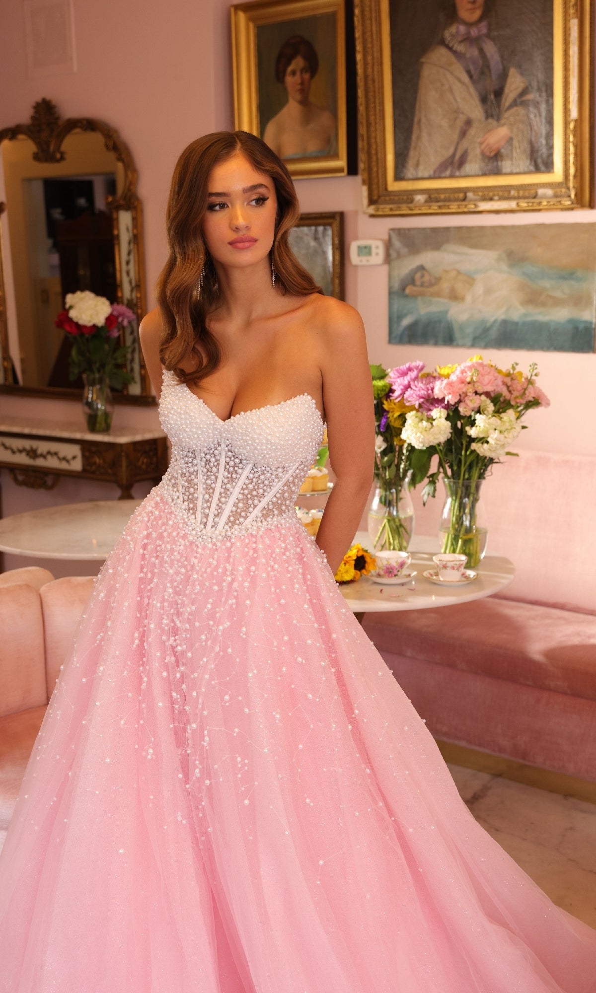 A woman in the Ava Presley 29226 Corset Tulle Strapless Ball Gown stands in an elegant room decorated with flowers, mirrors, and framed portraits.