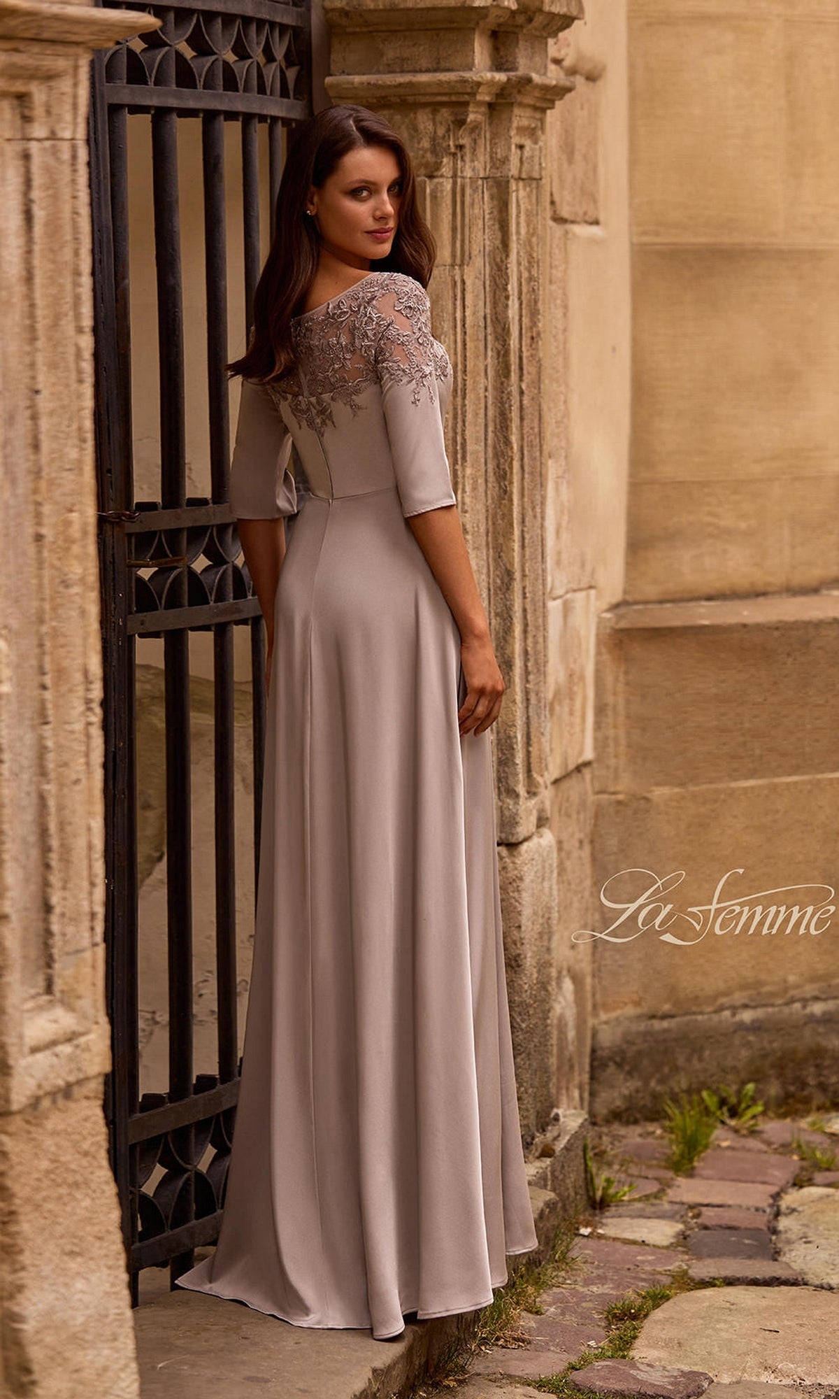 Woman in Long Formal Dress: La Femme 29227 with lace bodice and A-line silhouette stands by an old stone building and iron gate; “La Femme” logo is visible.
