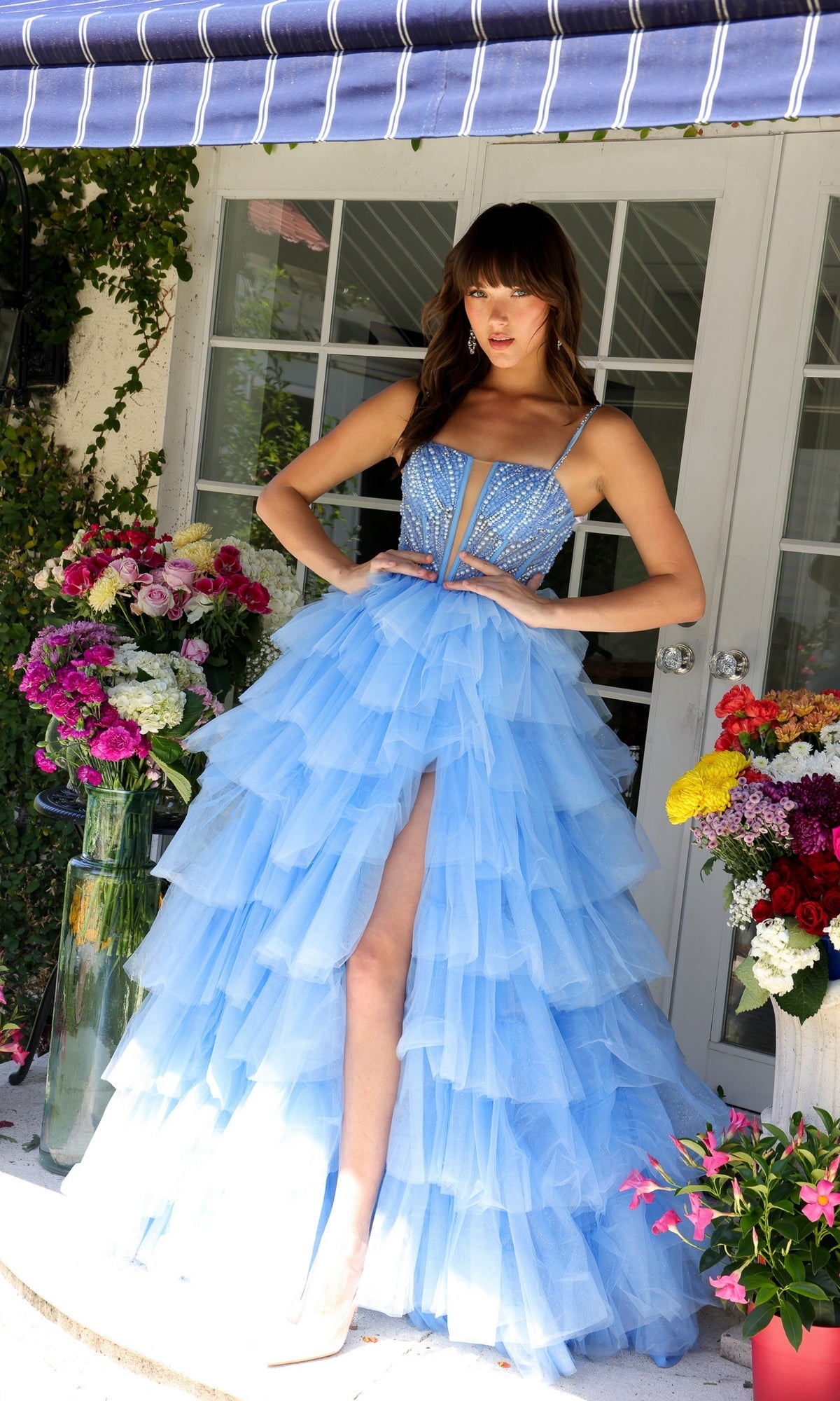 A woman wearing the Long Prom Dress 29232 by Ava Presley, an a-line ruffle ball gown with a sweetheart neckline, stands before glass doors surrounded by vibrant floral arrangements in vases.