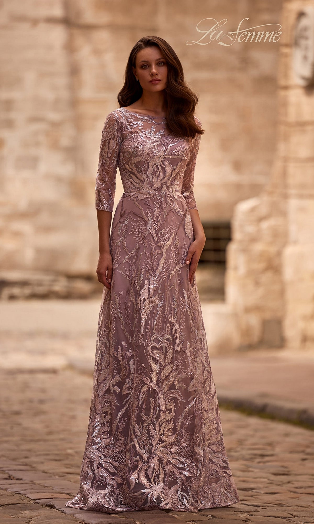 A woman in a mauve Long Formal Dress: La Femme 29233 with lace embroidery and three-quarter sleeves stands on a cobblestone street by a stone wall.