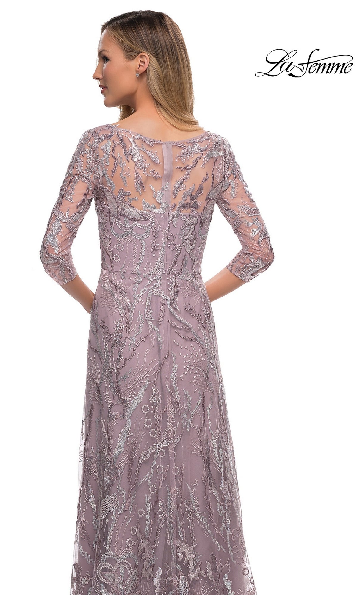 A woman wearing the Long Formal Dress: La Femme 29233, a lavender floor-length gown with silver lace and sheer sleeves, stands facing away against a white background.