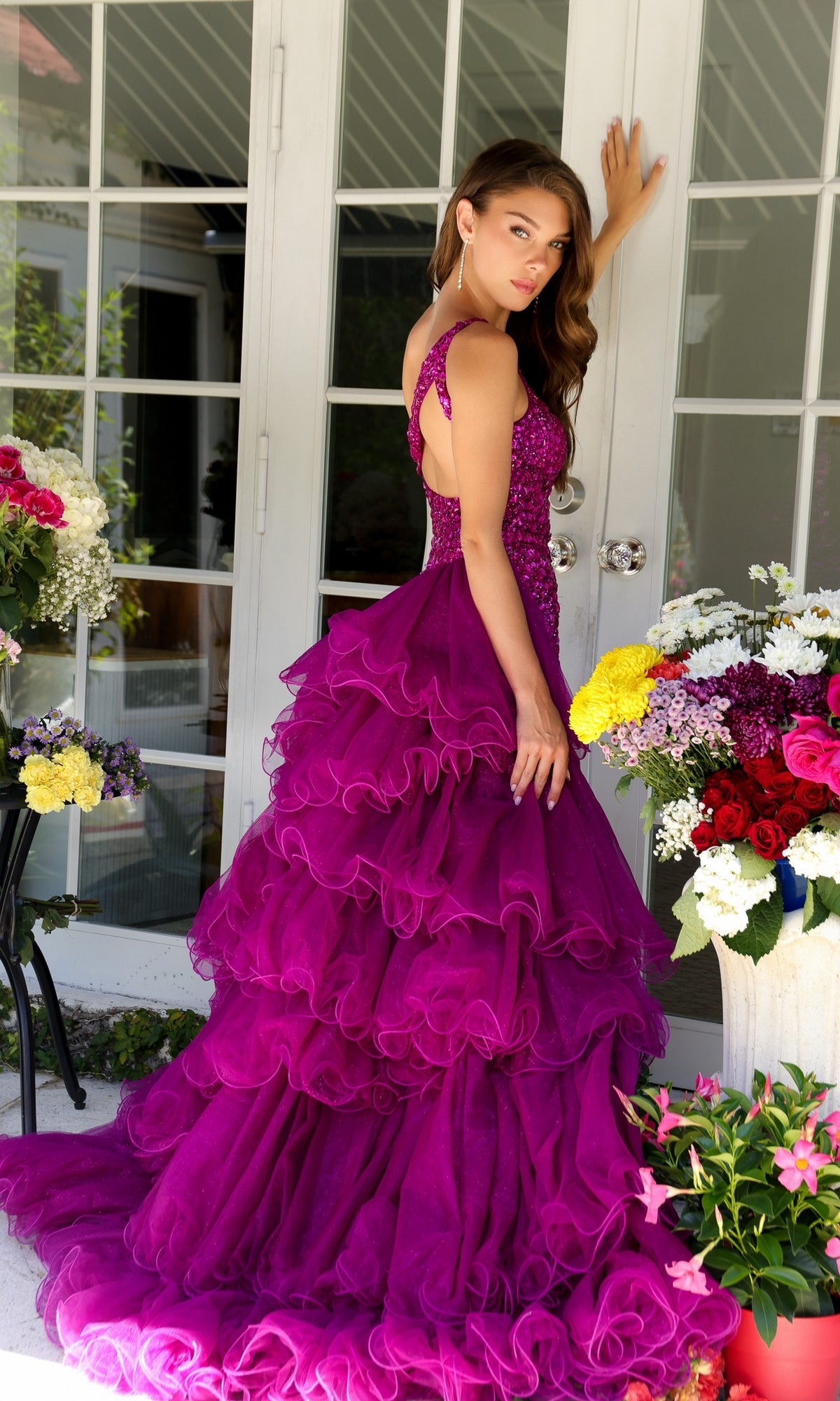 A woman in the Ava Presley 29236 One-Shoulder Beaded Prom Dress poses by glass doors, framed by potted flowers and plants. The tiered, ruffled magenta gown radiates elegance and charm.