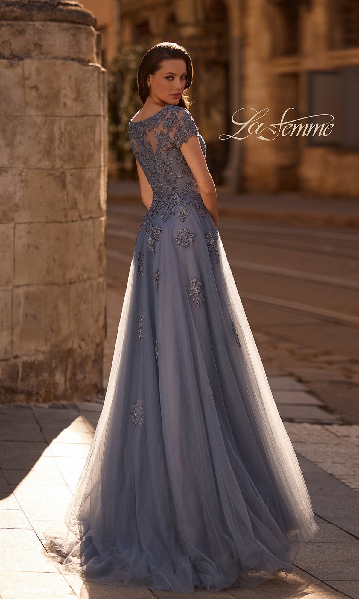 Woman in a Long Formal Dress: La Femme 29380 with lace appliques stands outdoors on a stone path, facing away.