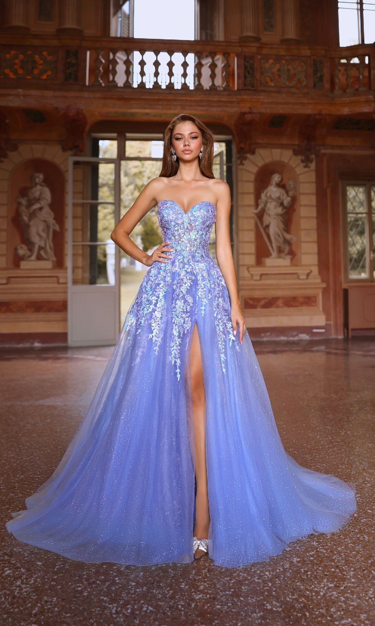 A woman in the Strapless Glitter Prom Ball Gown: Aerie Couture 293, featuring floral lace embroidery and a thigh-high slit, stands in an ornate room with large windows and classical statues.