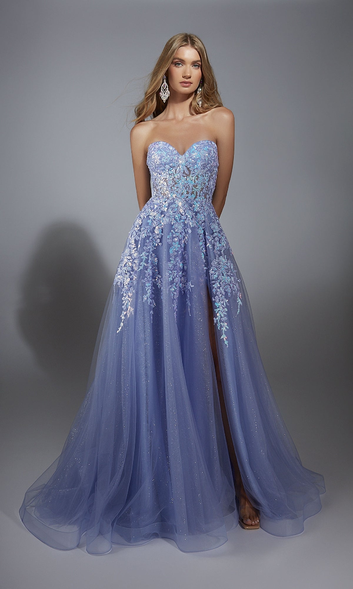 A woman in the Strapless Glitter Prom Ball Gown: Aerie Couture 293 stands against a plain gray background, showcasing its lavender hue with floral lace embroidery and a sheer overlay.