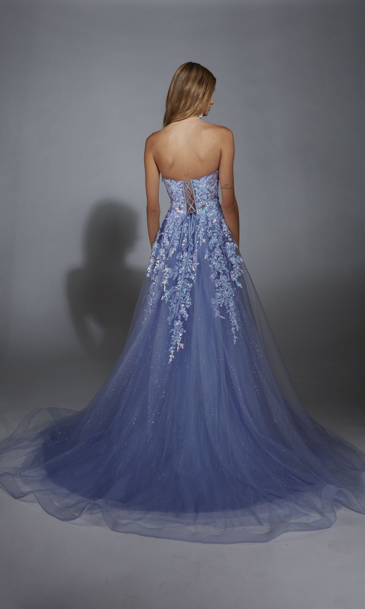 A woman wears the Strapless Glitter Prom Ball Gown: Aerie Couture 293, a strapless, floor-length blue dress with floral lace embroidery, standing with her back to a plain gray background.
