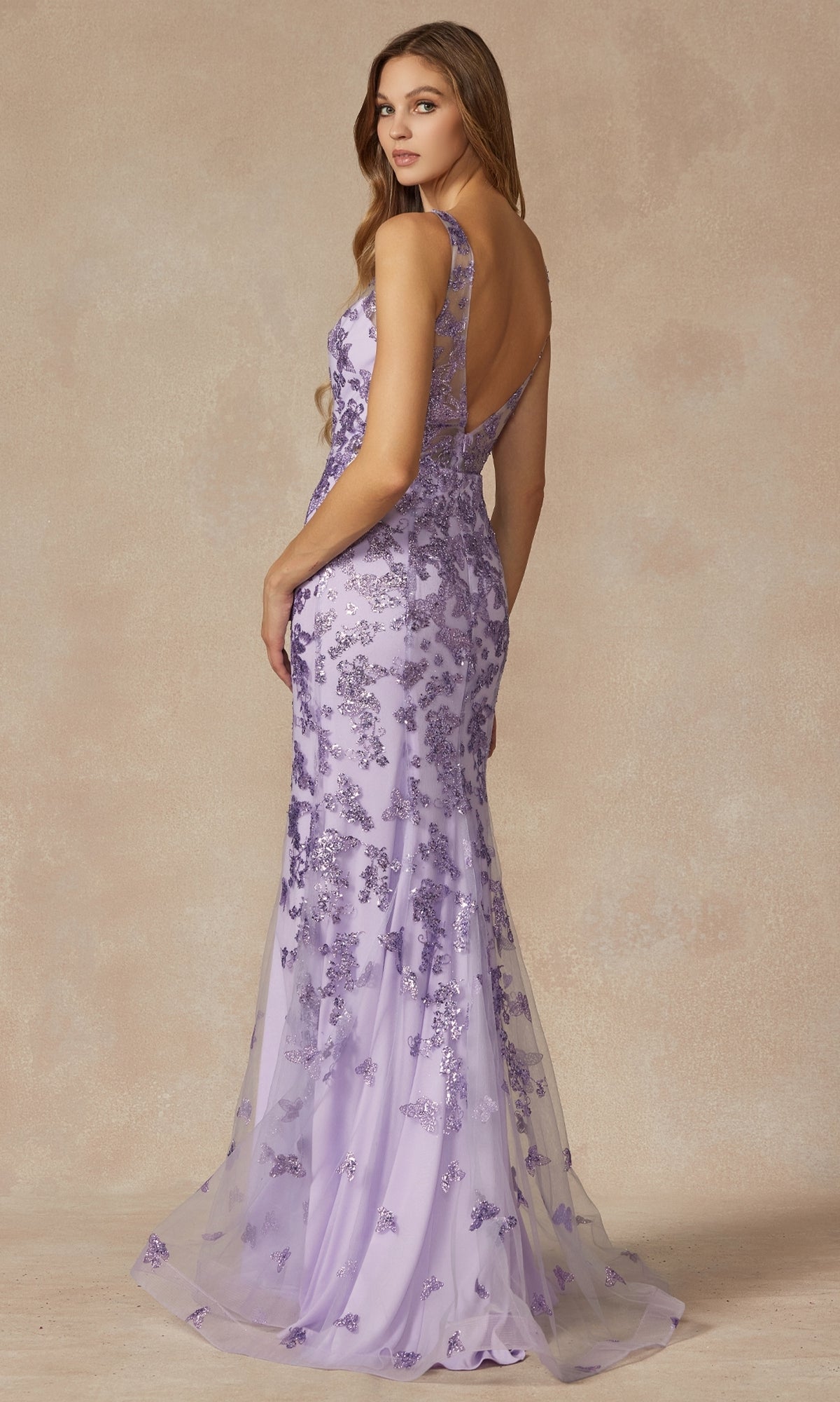 A woman models the Juliet 294 Glitter-Print Long Prom Dress, featuring a floor-length lavender design with floral lace embellishments and an open back, against a neutral background.