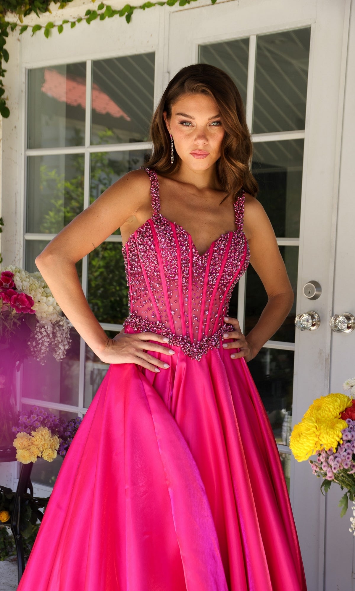 A woman stands before glass doors wearing the Long Formal Dress 29521 by Ava Presley—a bright pink, floor-length a-line gown with a beaded corset bodice and floral decorations in the background.