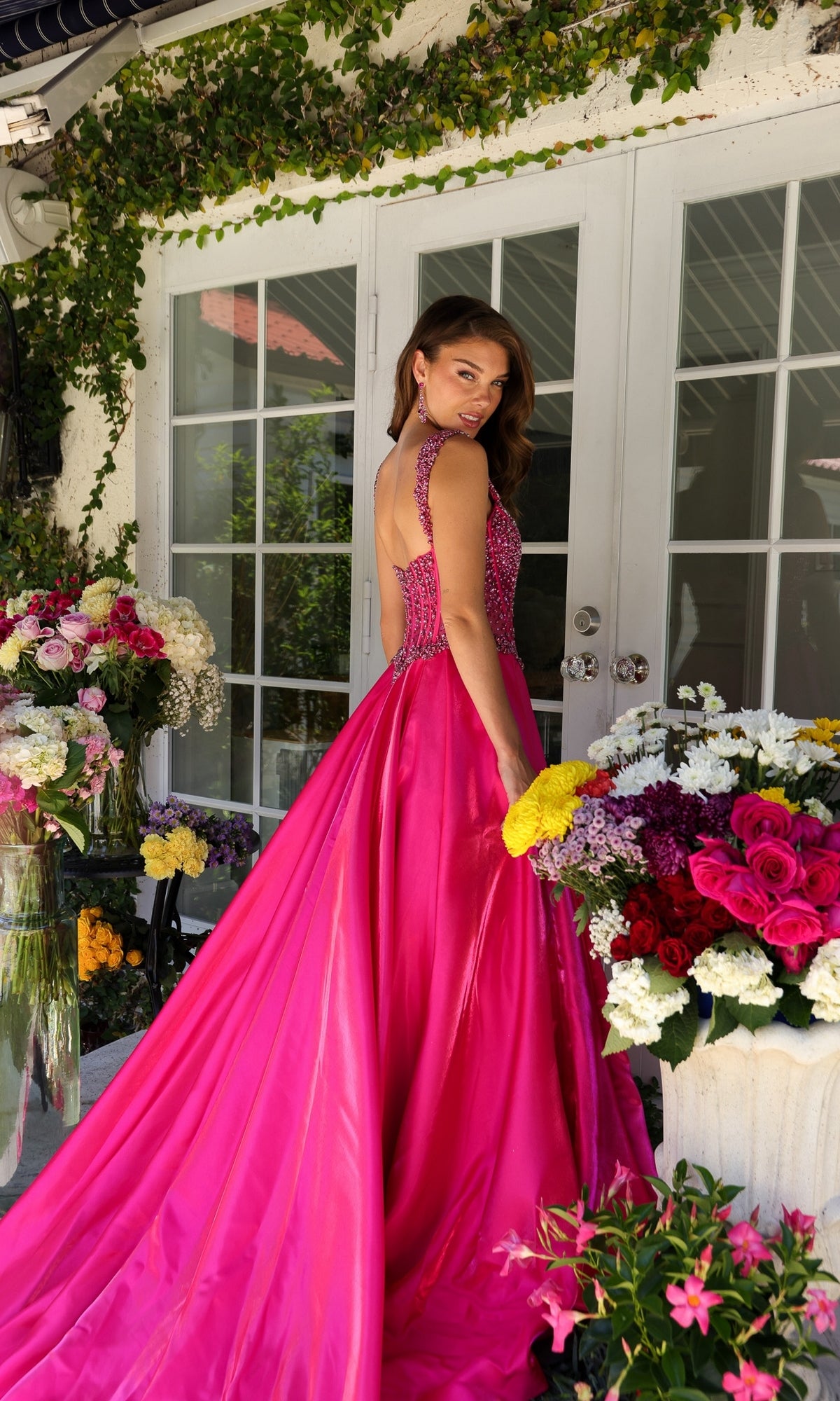 Wearing the Ava Presley Long Formal Dress 29521, a woman in a bright pink, floor-length A-line gown with a beaded corset bodice stands on a patio among colorful flower arrangements, looking over her shoulder at the camera.