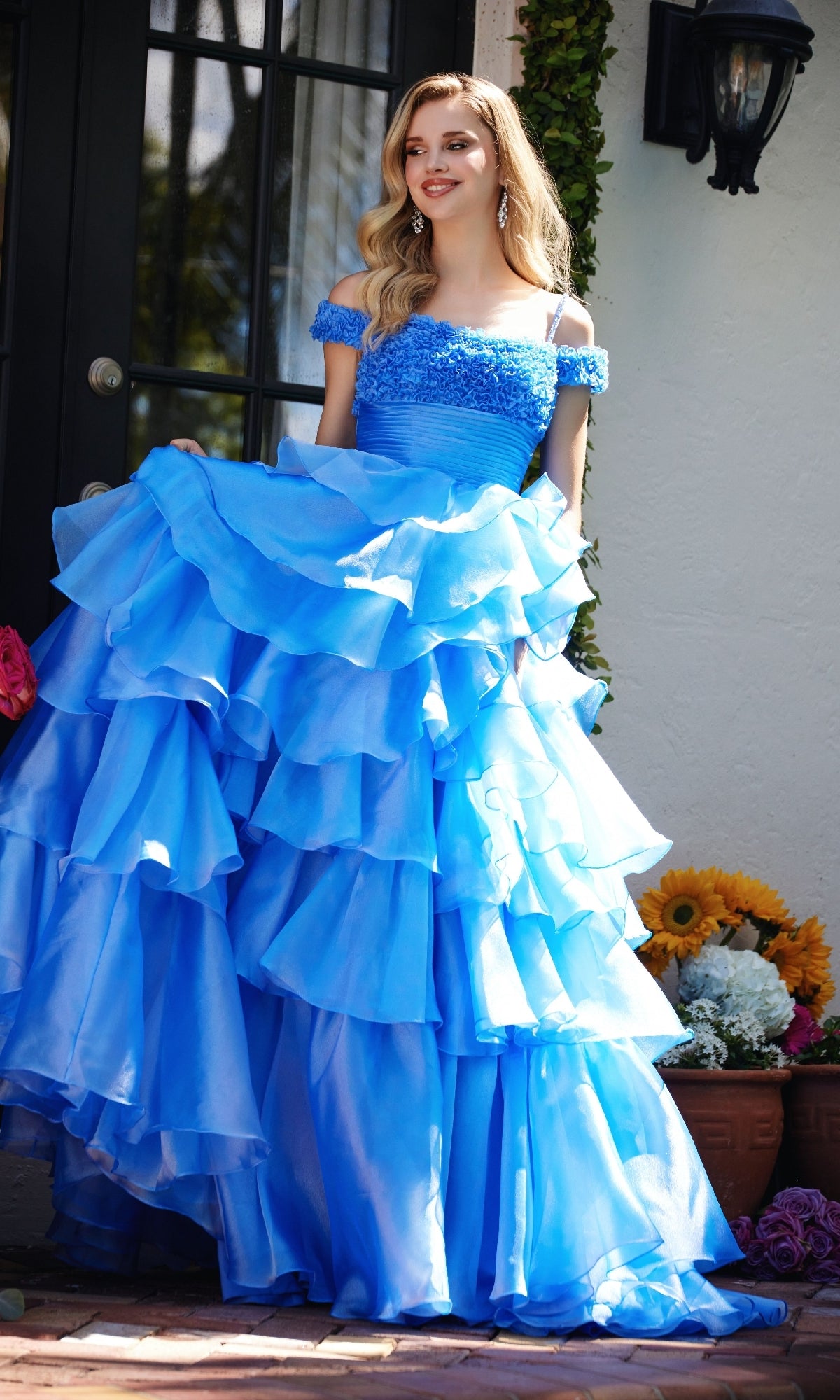 A woman stands outside in the Ava Presley 29544 Ruffled Prom Ball Gown, featuring a bright blue off-the-shoulder design and princess a-line skirt, holding her dress with sunflowers and potted plants nearby.
