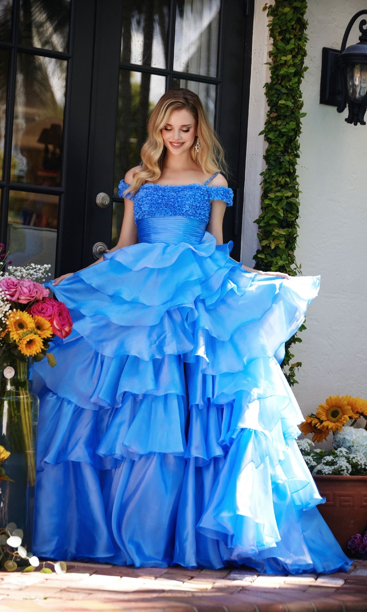 A woman wearing the Ava Presley 29544 ruffled blue prom ball gown with off-the-shoulder straps stands outside near glass doors, surrounded by sunflowers and colorful flowers.