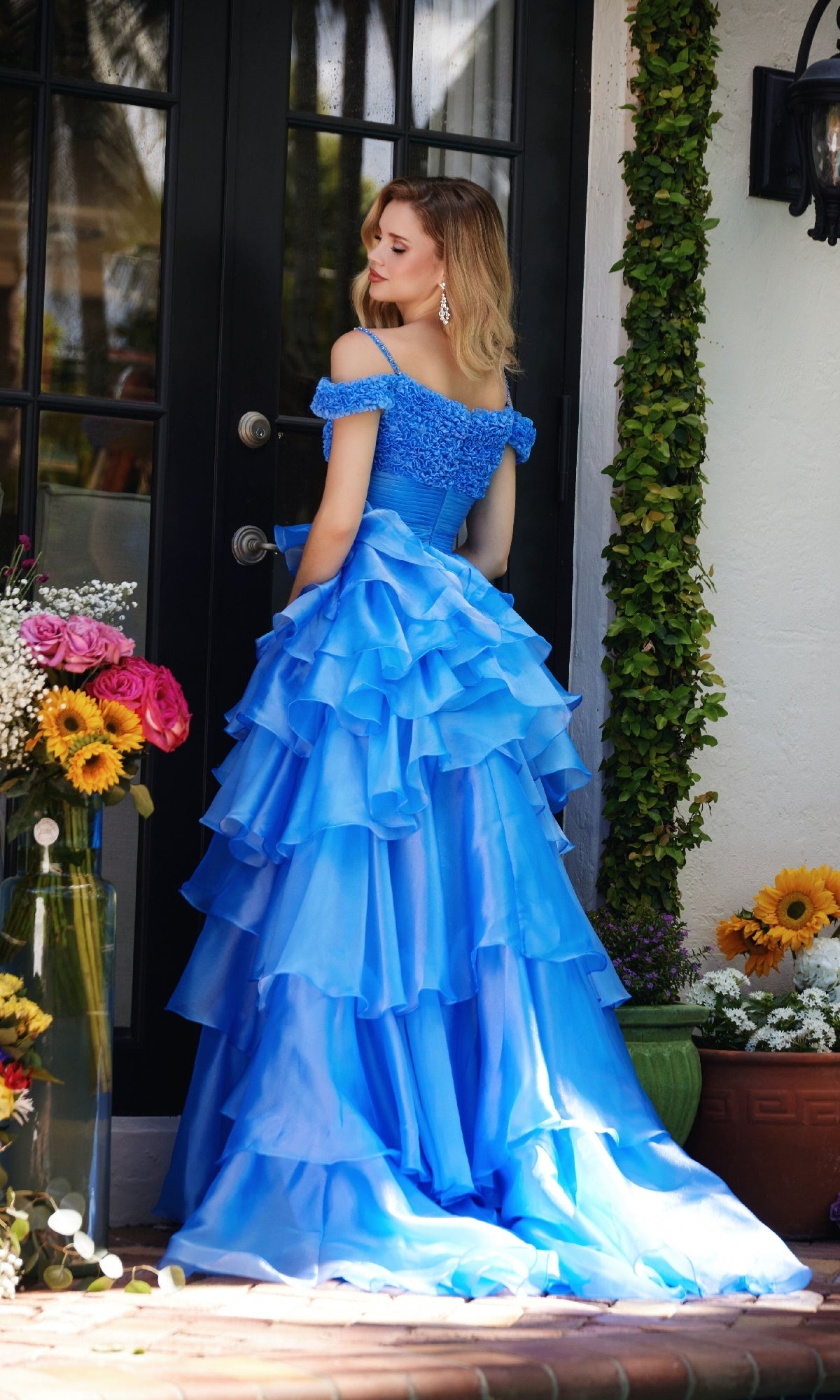 A woman in the Ava Presley 29544 Ruffled Prom Ball Gown with off-the-shoulder straps and a princess A-line skirt stands outside by glass doors, surrounded by flowers and greenery.