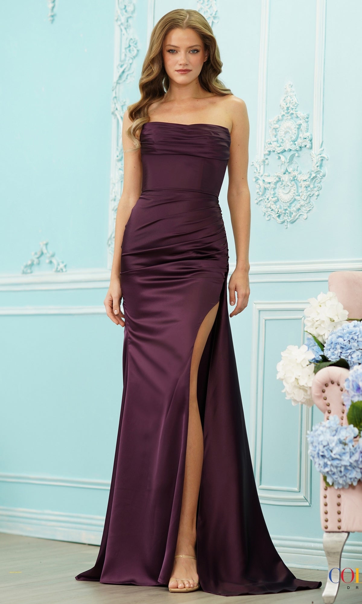 A woman in the Strappy-Back Strapless Long Satin Prom Dress 2968 stands in a light blue ornate room near a pink chair and white and blue flowers.