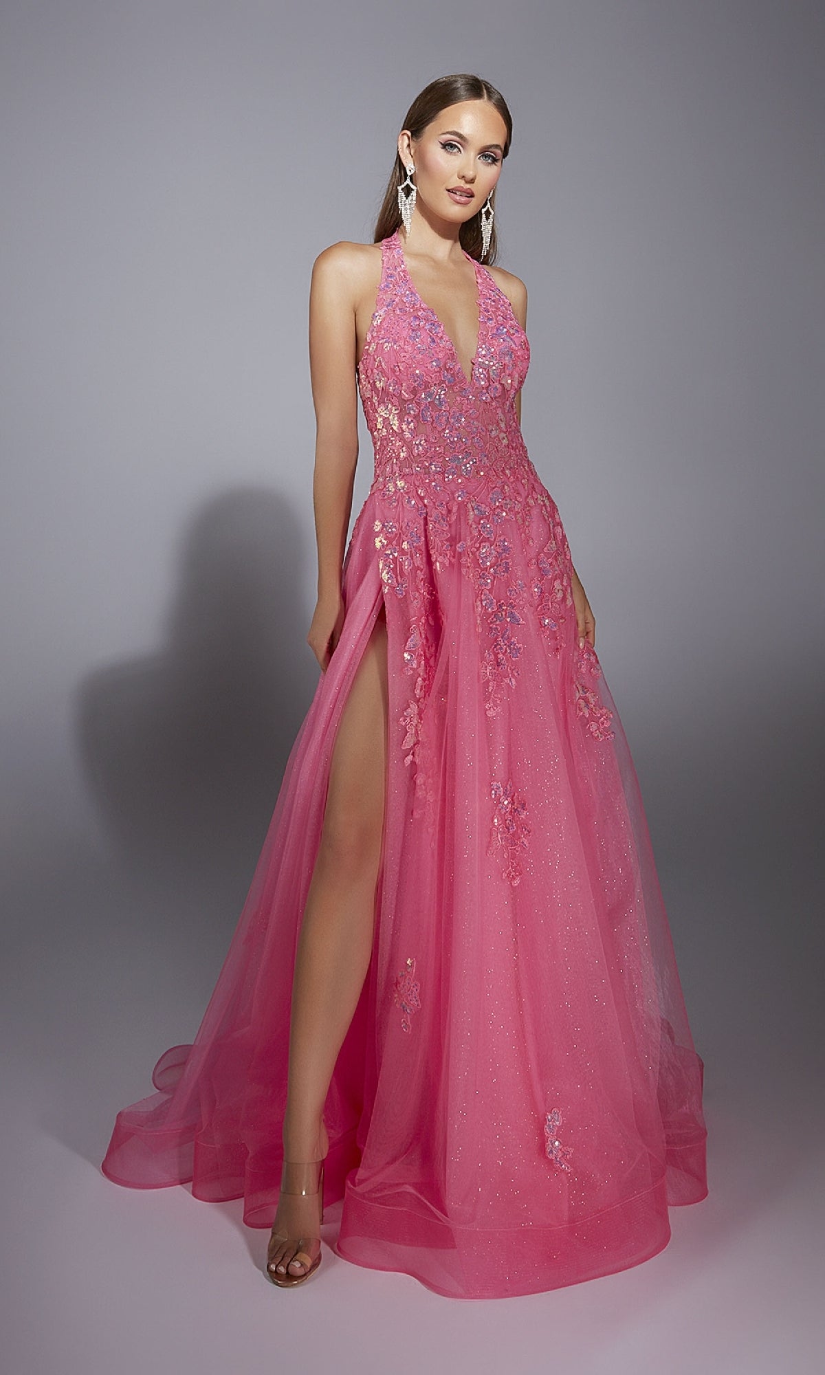 A woman poses against a plain gray background wearing the Long Halter Prom Dress: Aerie Couture 296—a pink, sparkly gown with floral embellishments, lace embroidery, a deep V neckline, and a high slit.