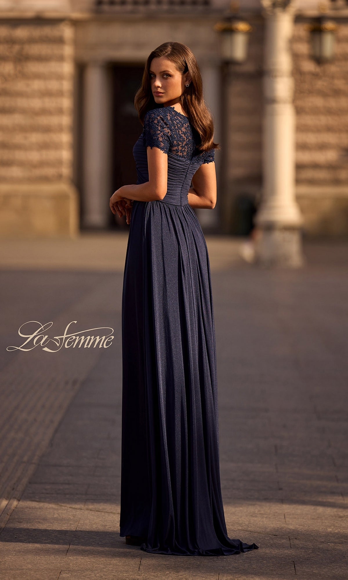 Woman wearing the Long Formal Dress: La Femme 29772 in navy blue, featuring a ruched bodice and lace sleeves, stands outdoors on a stone pathway, facing away with head turned.