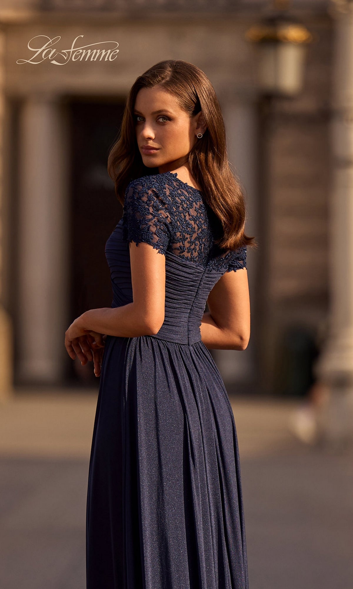 A woman in the Long Formal Dress: La Femme 29772 poses outdoors in a navy blue lace gown with short sleeves and a ruched fitted bodice, with blurred architecture behind her.