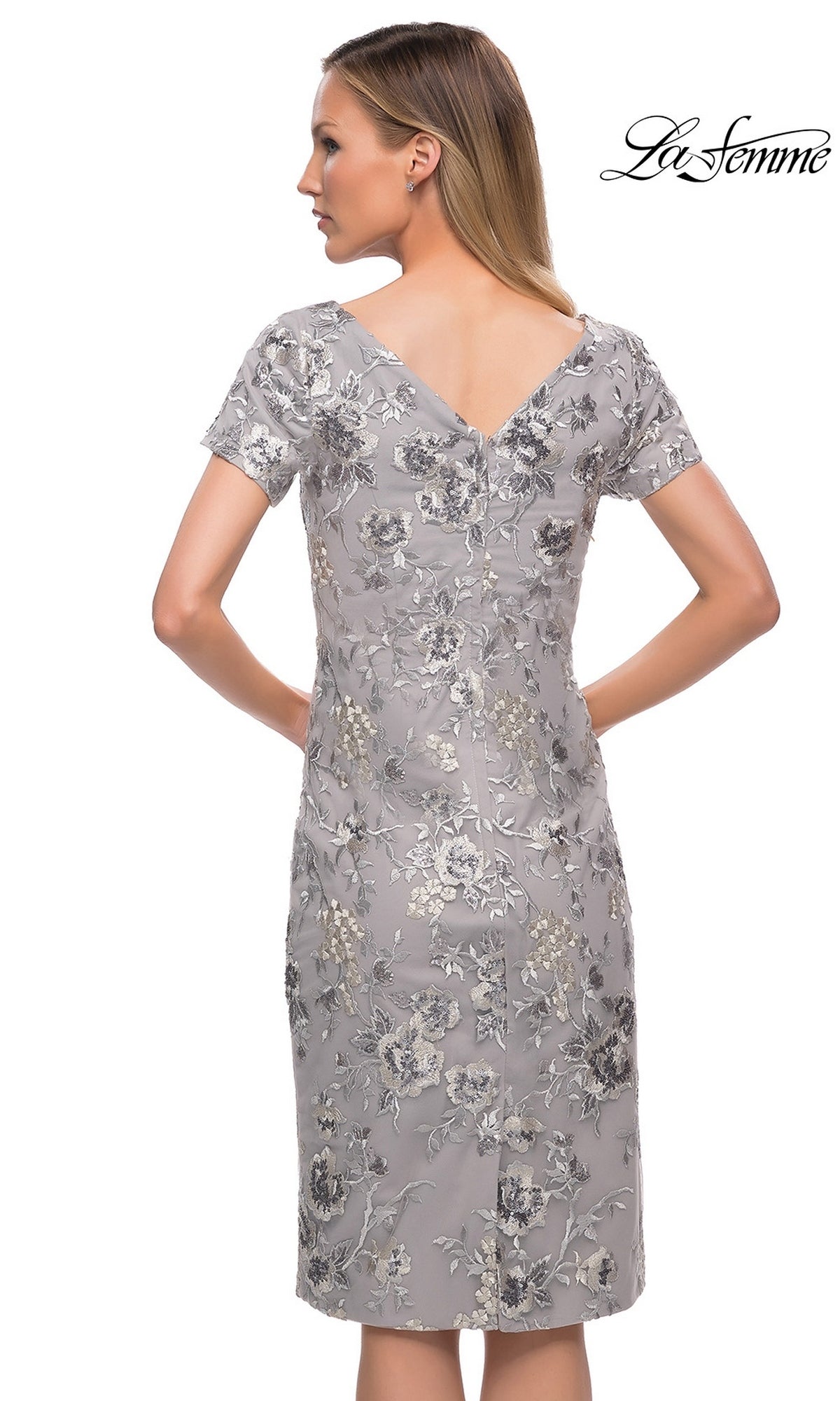 A woman with straight blonde hair is seen from the back wearing the Knee Length Wedding Guest Dress: La Femme 29824 in silver floral-lace embroidery, logo visible.