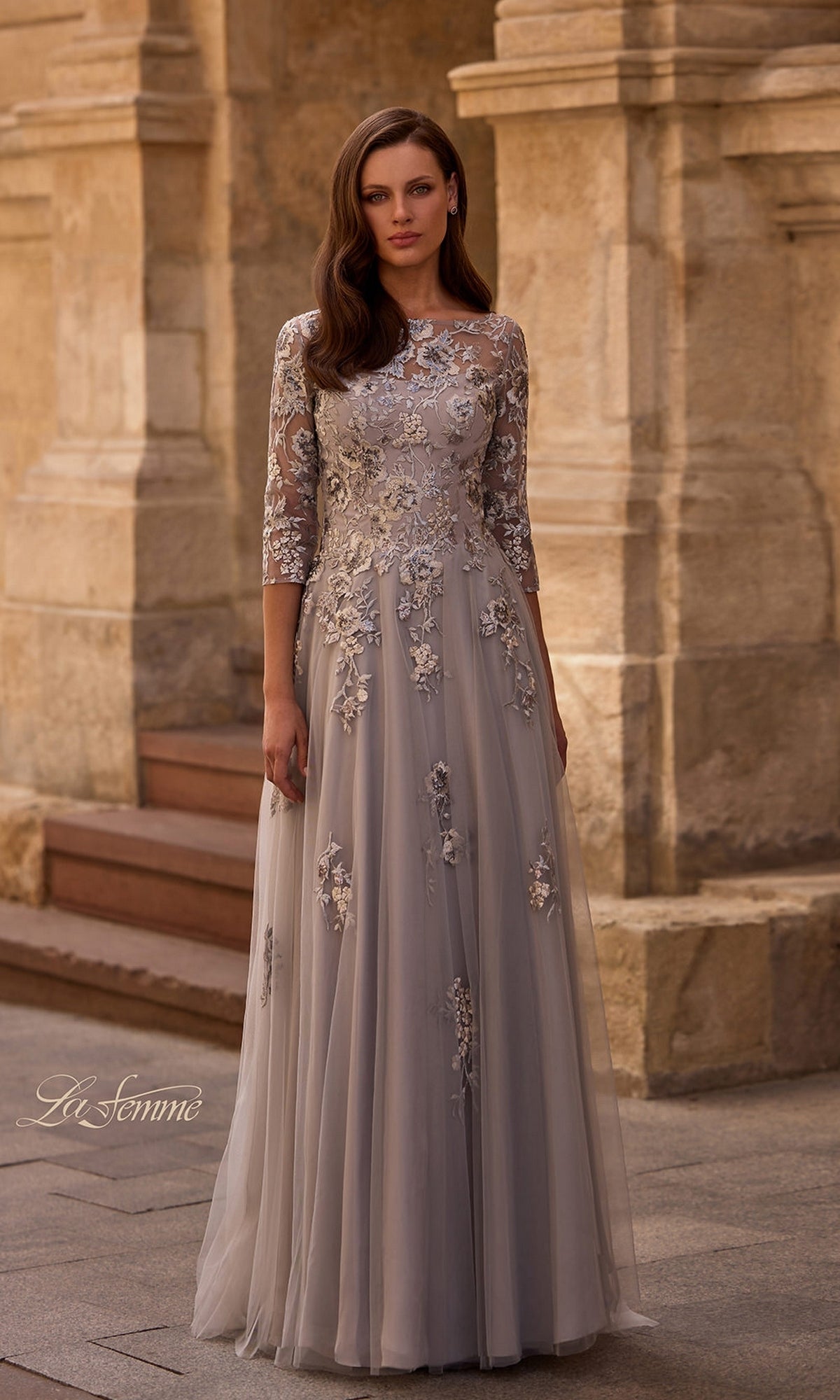 Woman stands outside in a silver Long Formal Dress: La Femme 29825 with lace floral embroidery and three-quarter sleeves. Stone building in the background.