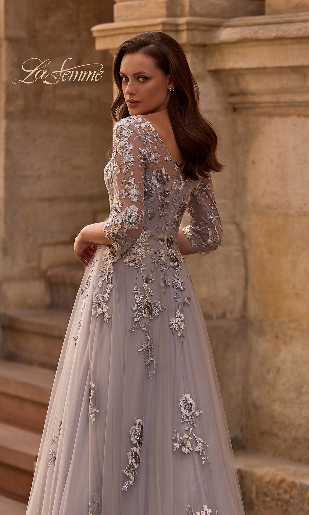 Wearing the Long Formal Dress: La Femme 29825, a woman in a silver floral embroidered tulle gown stands by stone steps, glancing over her shoulder.