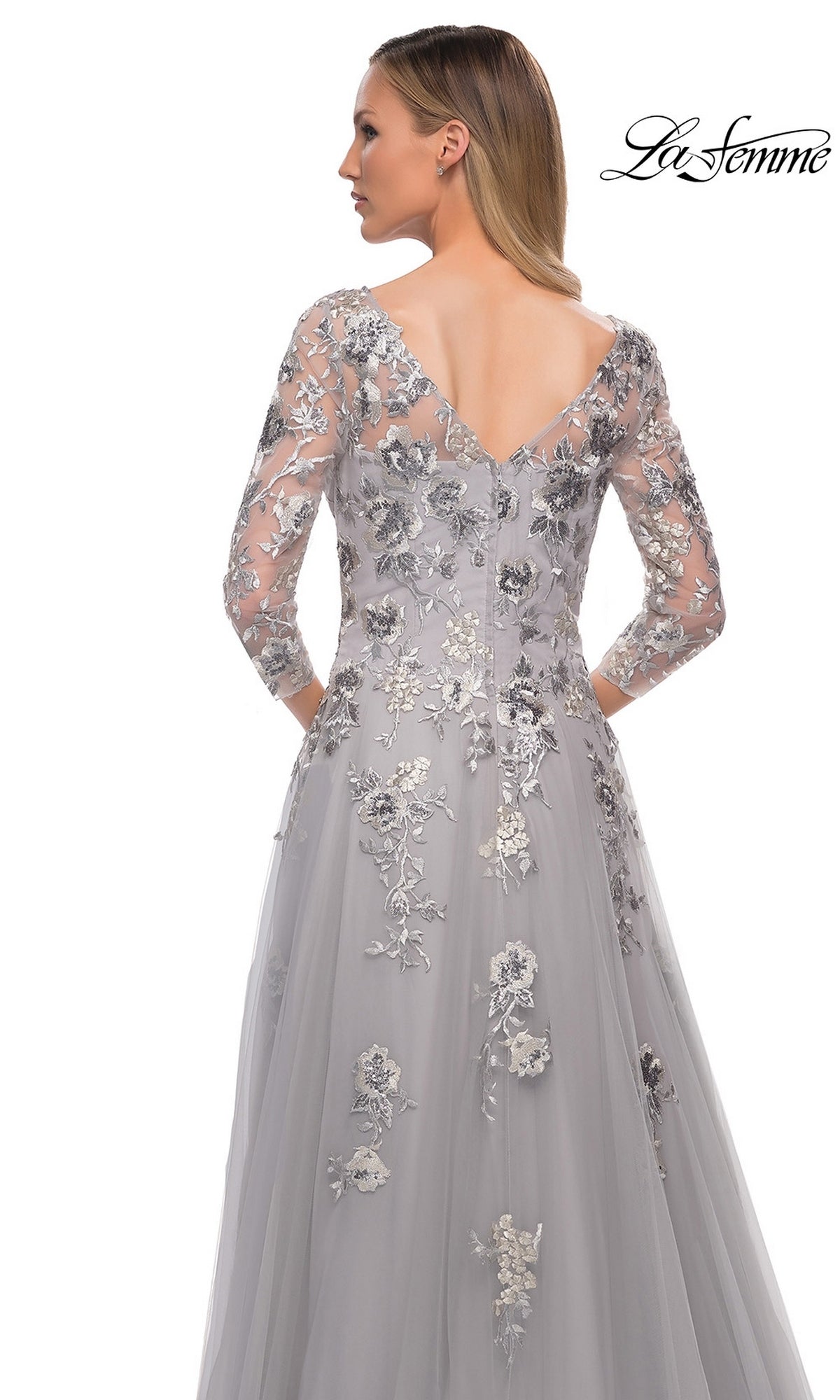 Woman in Long Formal Dress: La Femme 29825—silver tulle gown with floral embroidery, sheer sleeves, V-back; hands behind her back. La Femme logo in corner.
