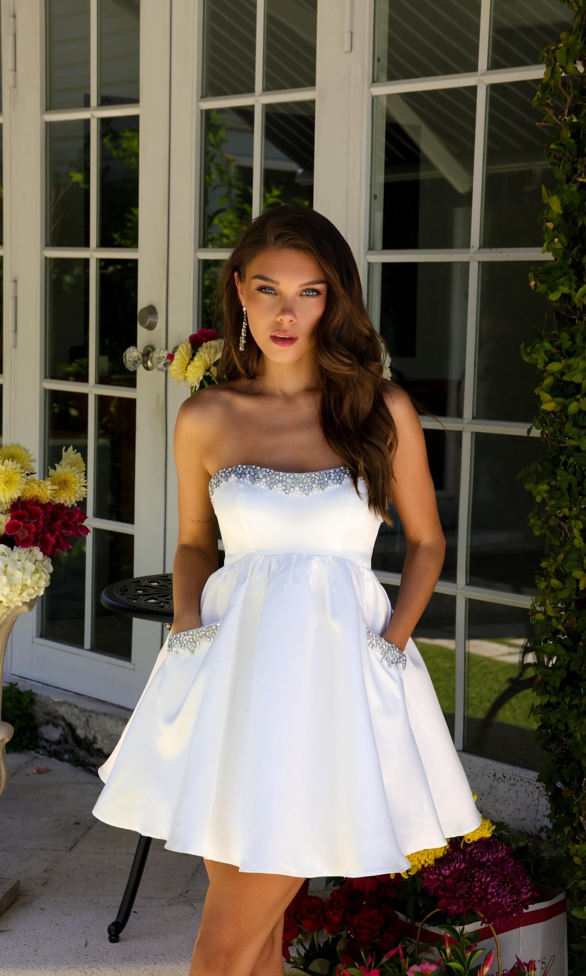 A woman stands outdoors by glass doors, surrounded by flowers, wearing the Ava Presley 29837 Strapless Short Hoco Dress with jeweled trim and pockets.