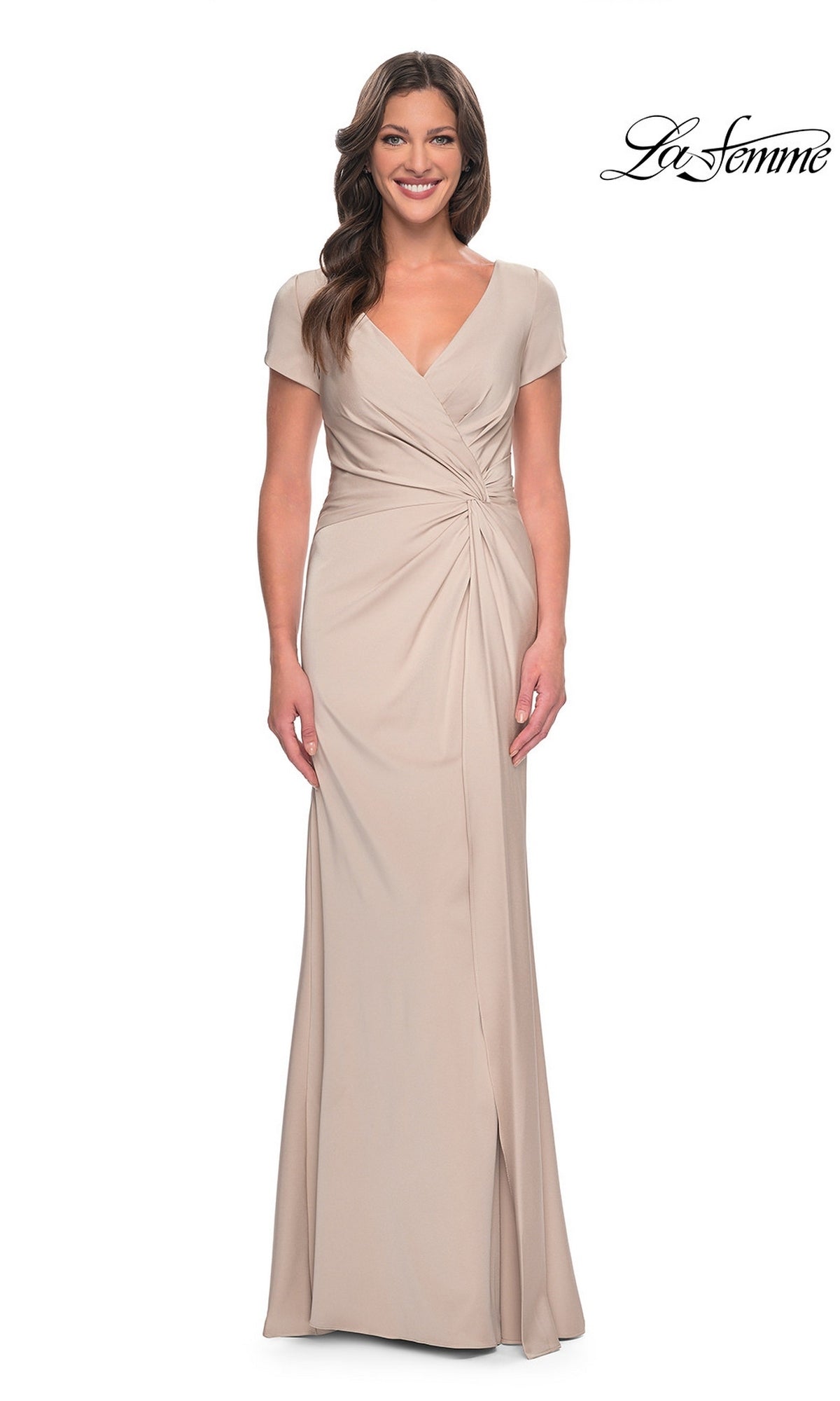A woman wearing a Long Formal Dress: La Femme 29926, featuring a floor-length, beige V-neck gown with short sleeves and a twist-knot waist detail.