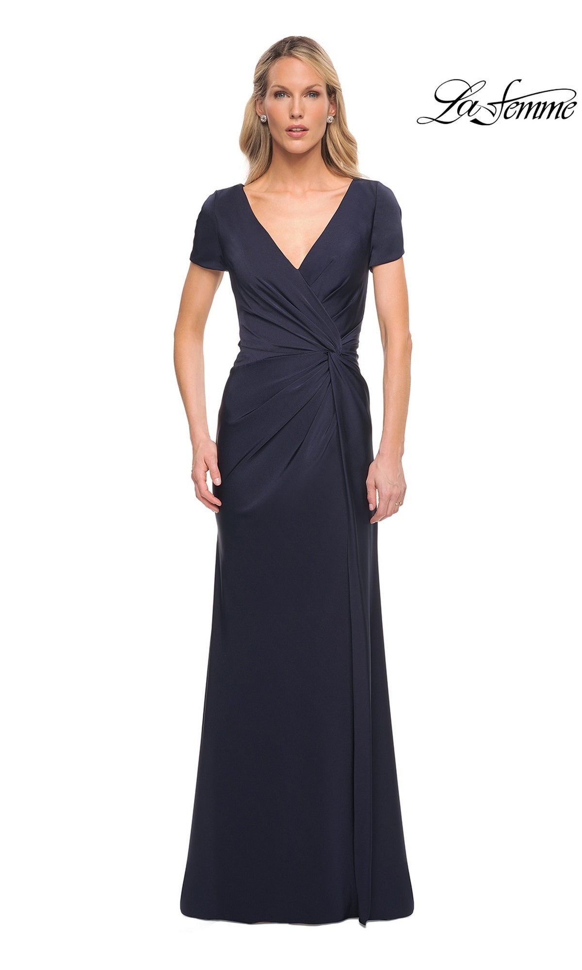 A woman wears a navy Long Formal Dress: La Femme 29926 featuring a V-neckline, short sleeves, and a draped, twist-knot waist.