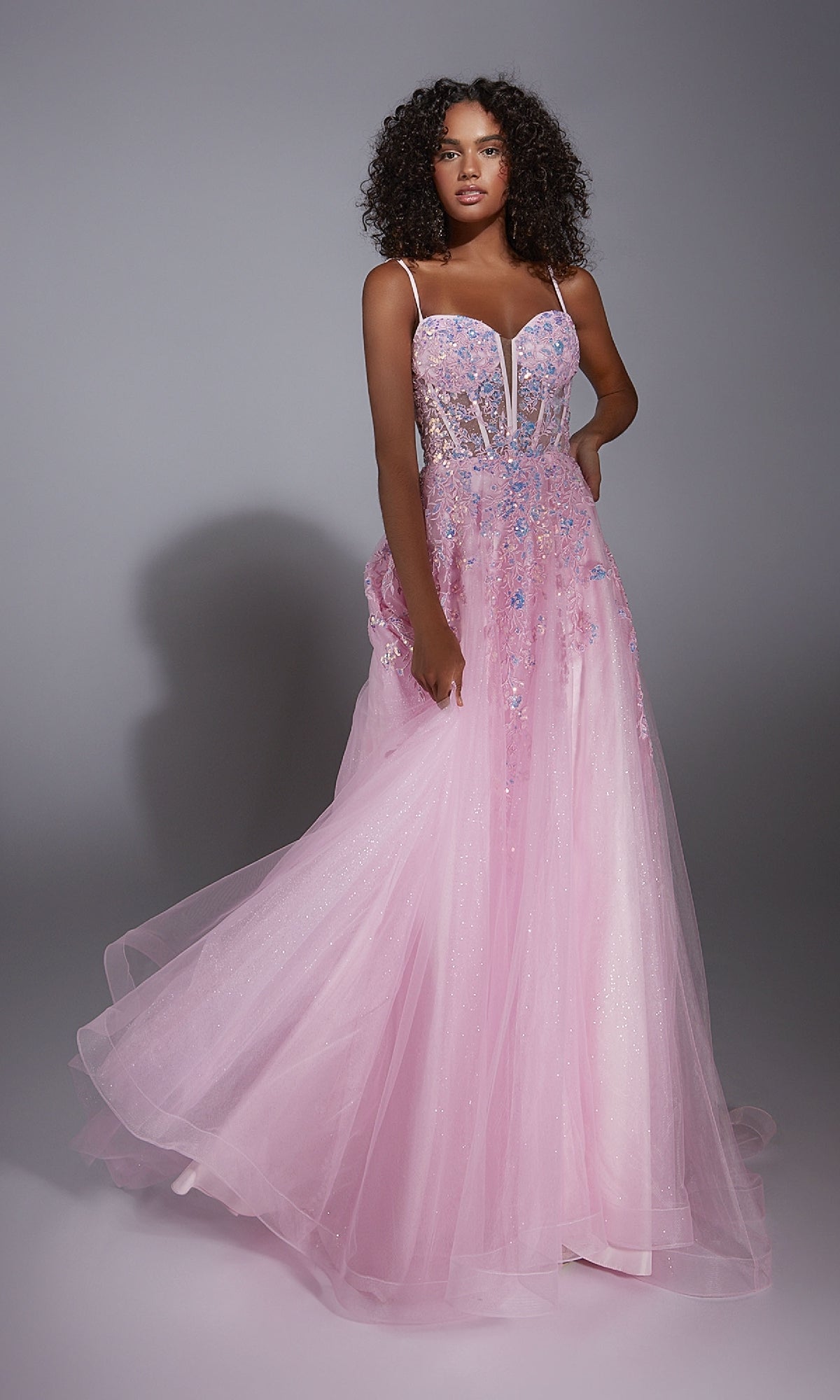 A woman stands in a studio wearing the Aerie Couture 299, a floor-length pink glitter tulle prom dress with thin straps, floral embellishments, and a fitted bodice.