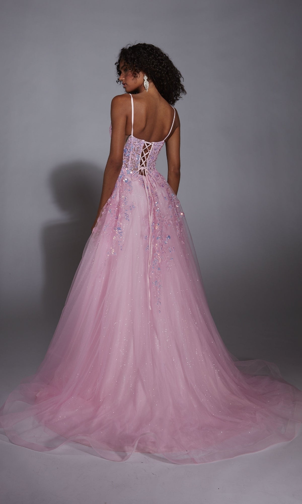 A woman faces away in the Aerie Couture 299, a floor-length light pink glitter tulle prom dress featuring thin straps, a lace-up back, and a sheer sparkly overlay.