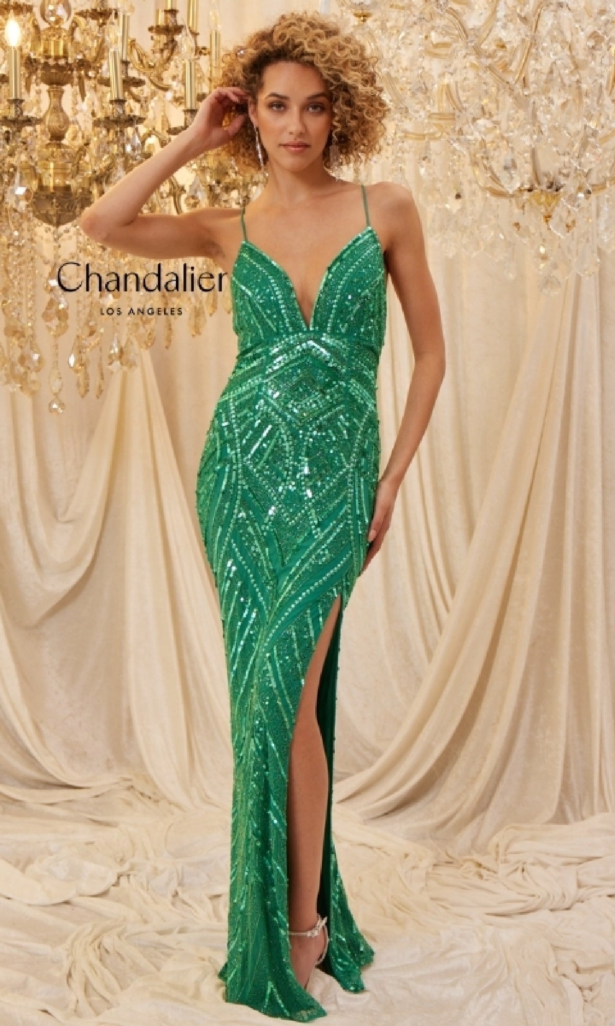 A woman wearing the Long Prom Dress: Chandalier 30023, featuring a form-fitting sequin design with a thigh-high slit and lace-up open back, stands before draped fabric and a crystal chandelier.