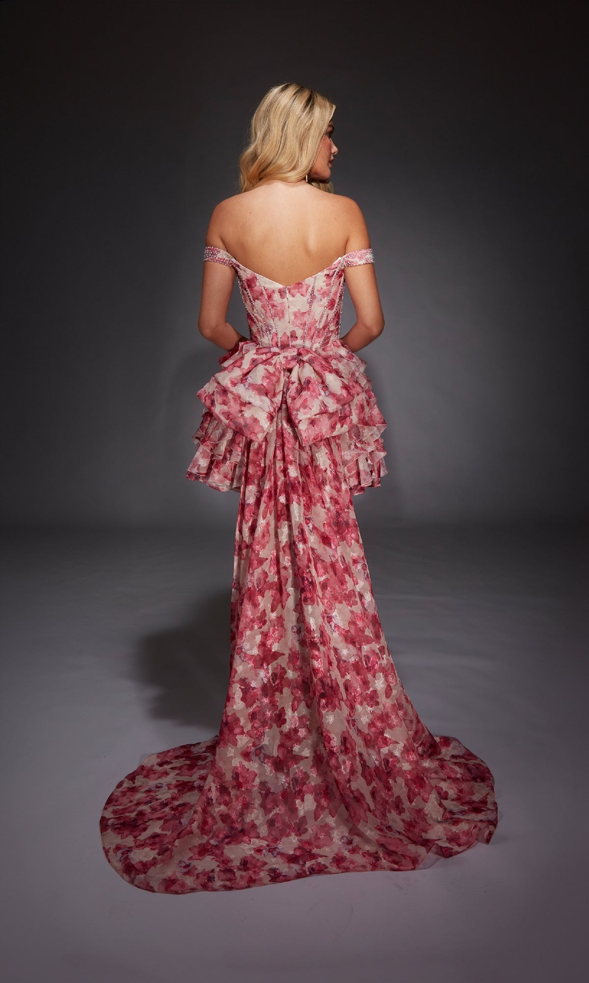 A woman stands facing away against a dark background, wearing the Alyce 30034 short homecoming dress—an off-the-shoulder, pink and white floral fit-and-flare style with ruffled layers.