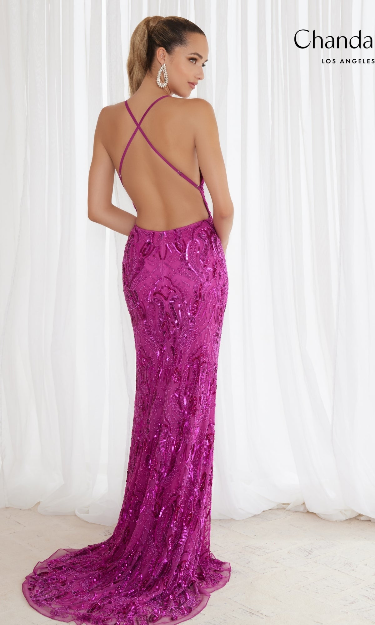 A woman faces away from the camera in front of white curtains, wearing the Long Prom Dress 30047 by Chandalier—a magenta sequin gown with a low, open back and crisscross straps.