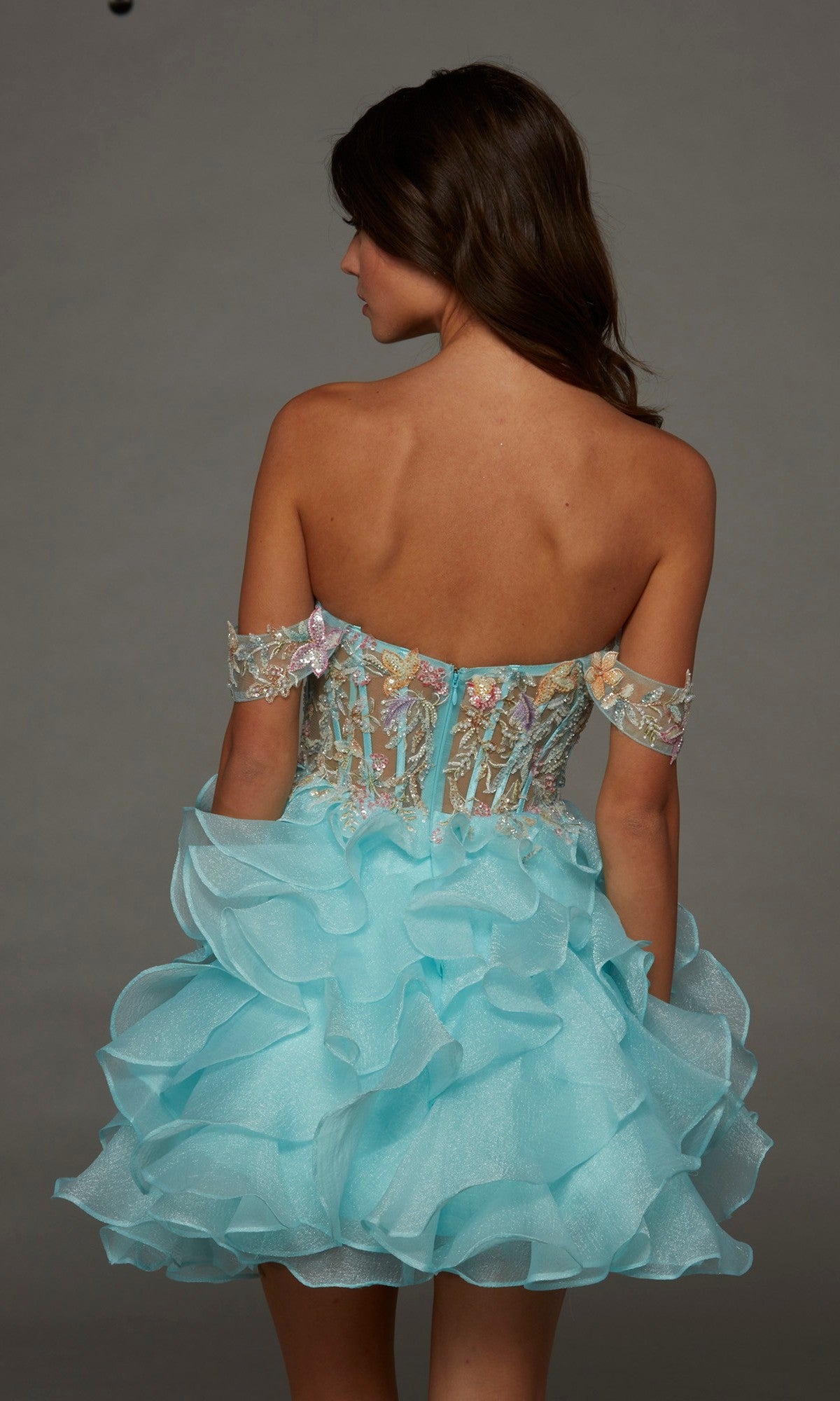 A woman stands with her back to the camera, wearing the Alyce 30087 Short Homecoming Dress—a strapless blue fit-and-flare style featuring ruffled details, beaded floral appliques, and off-the-shoulder sleeves—against a plain background.