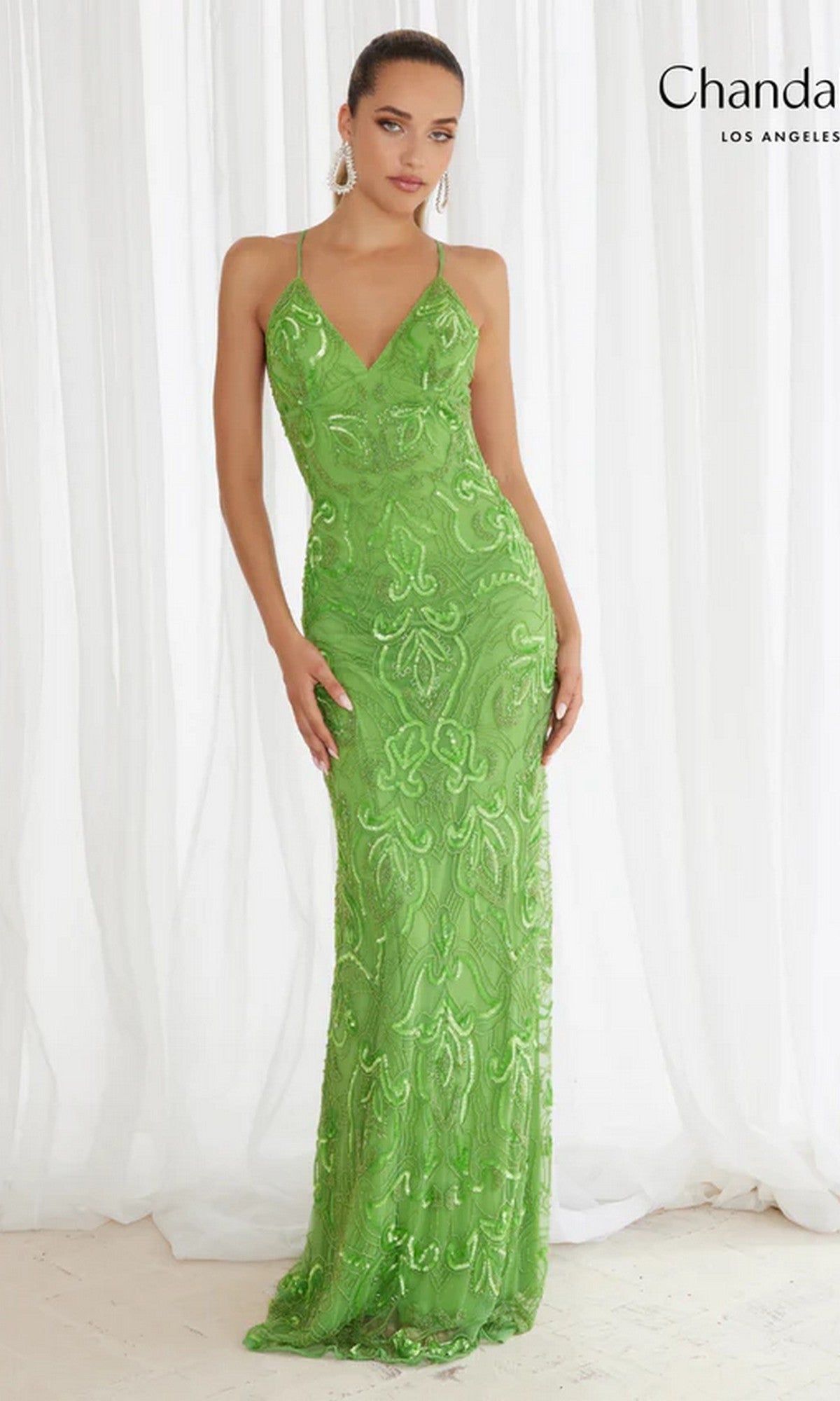A woman in the Sequin-Print Long Prom Dress: Chandalier 30087, a fitted, floor-length green gown with sequin detailing, stands before white curtains.