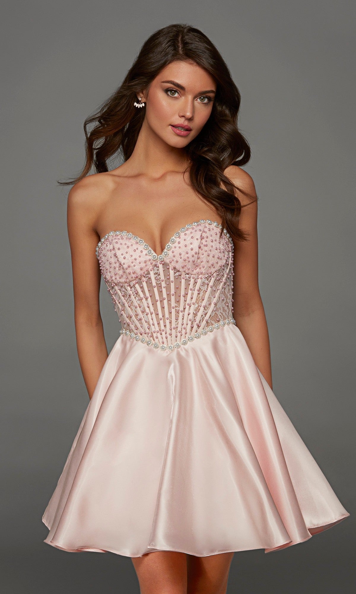 A woman stands against a plain gray background wearing the Alyce 30088 short homecoming dress in light pink, which features a strapless design with a beaded, semi-sheer corset bodice and satin skirt.